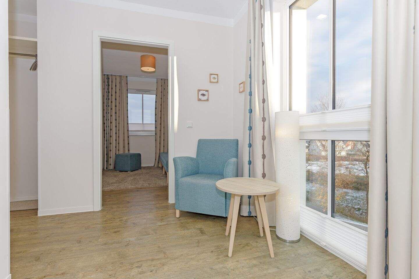 Ein Raum mit blauem Sessel, runden Tisch und Fensterblick. Im Hintergrund ist ein weiterer Raum sichtbar.