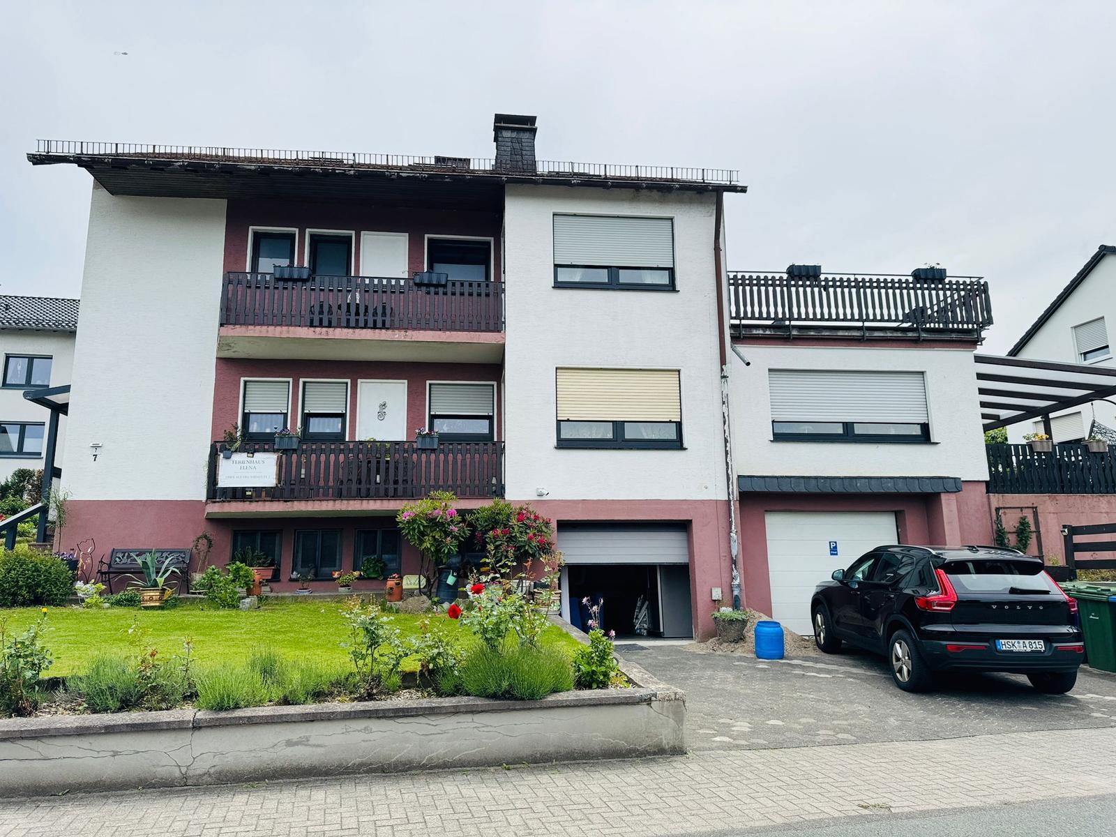 Zweistöckiges Haus mit Balkonen, Garage und Garten. Ein Auto parkt vor dem Haus.