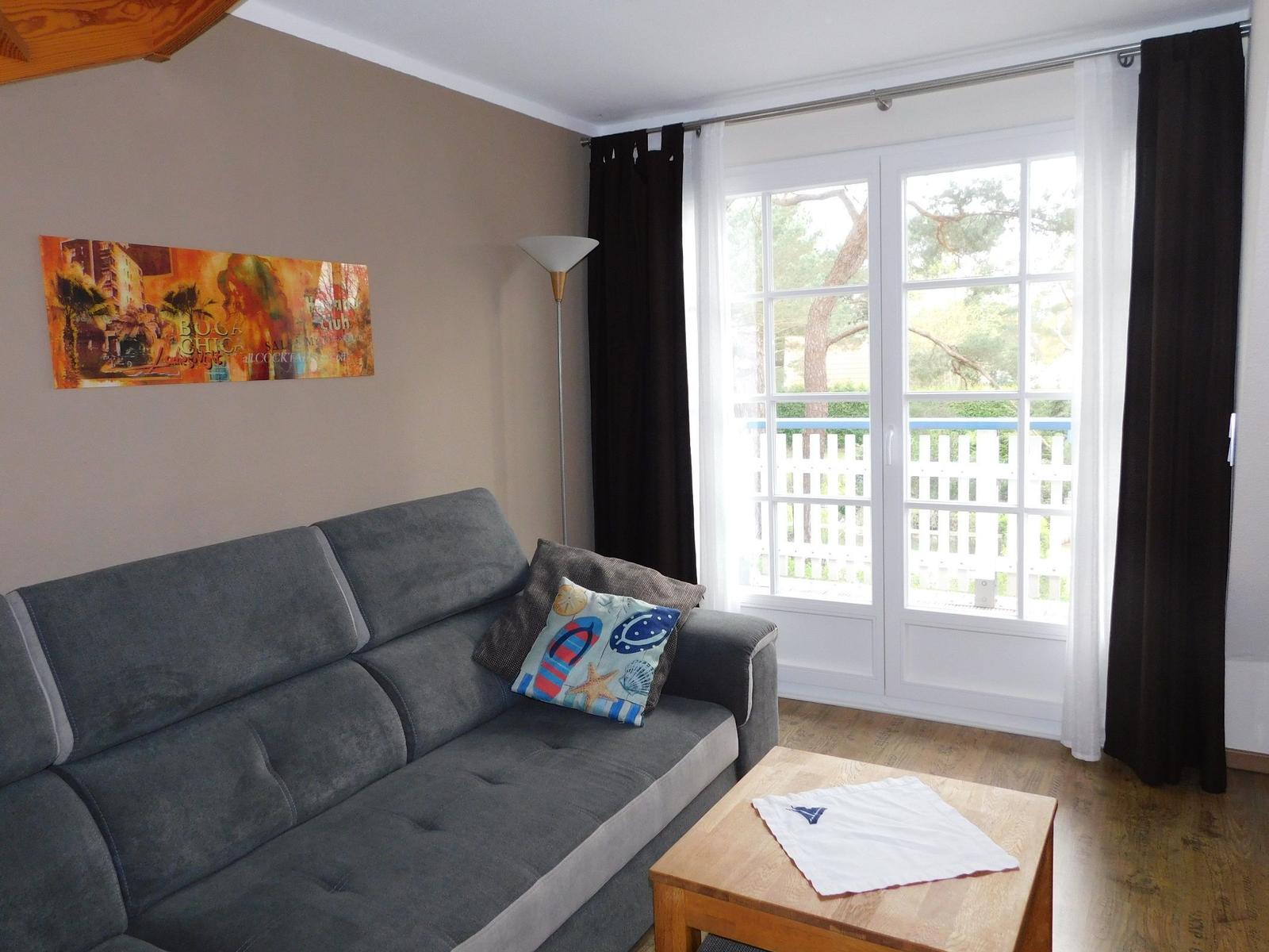 Ein Wohnzimmer mit grauem Sofa, Holztisch und Fenster mit Blick auf Balkon.