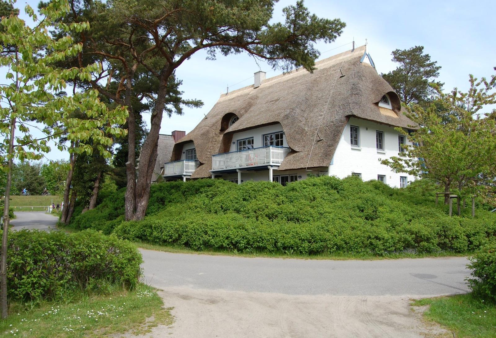 Haus mit Strohdach und Balkon, umgeben von Bäumen und Sträuchern.