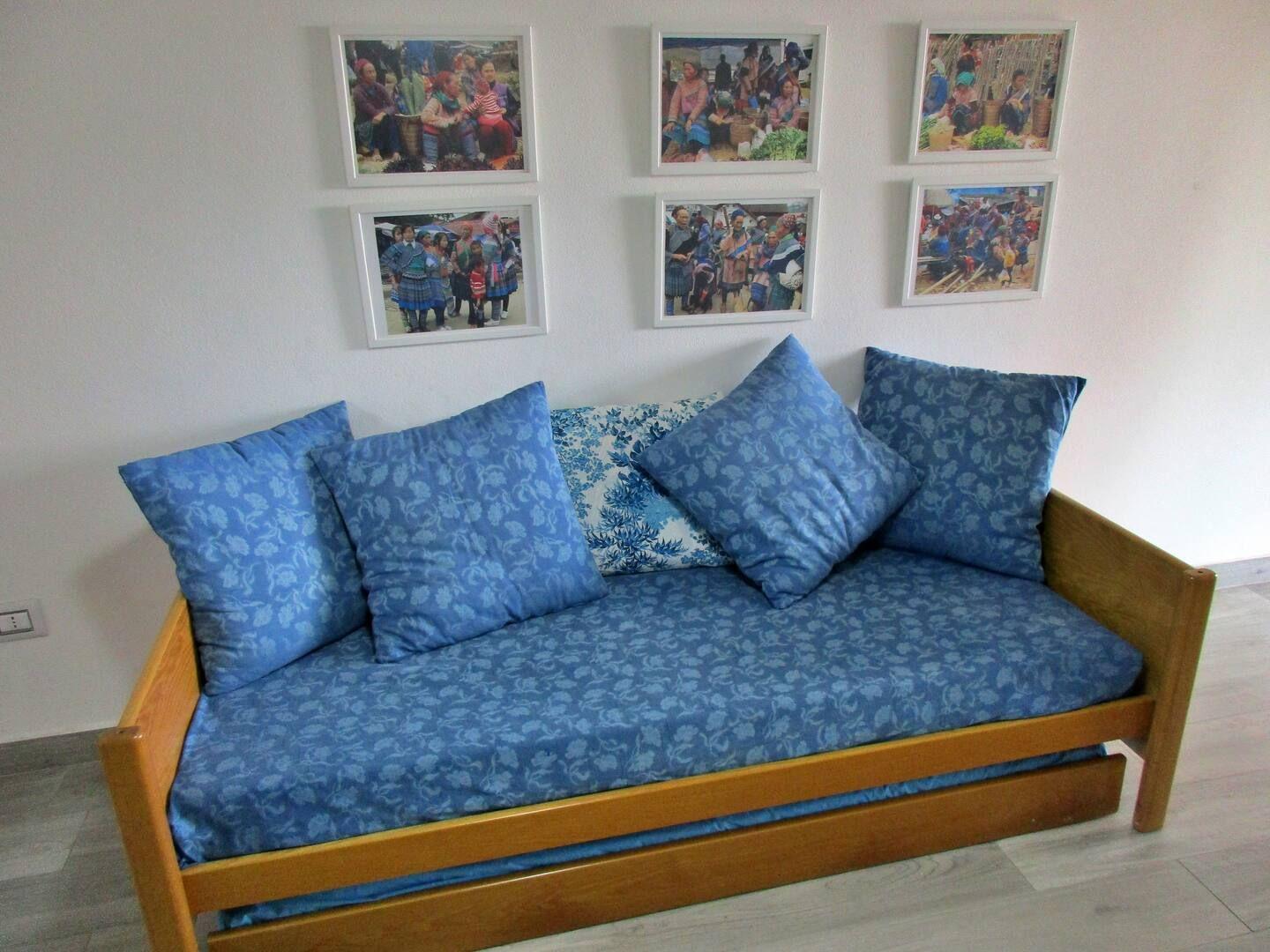 Ein Sofa mit blauen Kissen und einem Holzrahmen. An der Wand hängen sechs gerahmte Fotos.