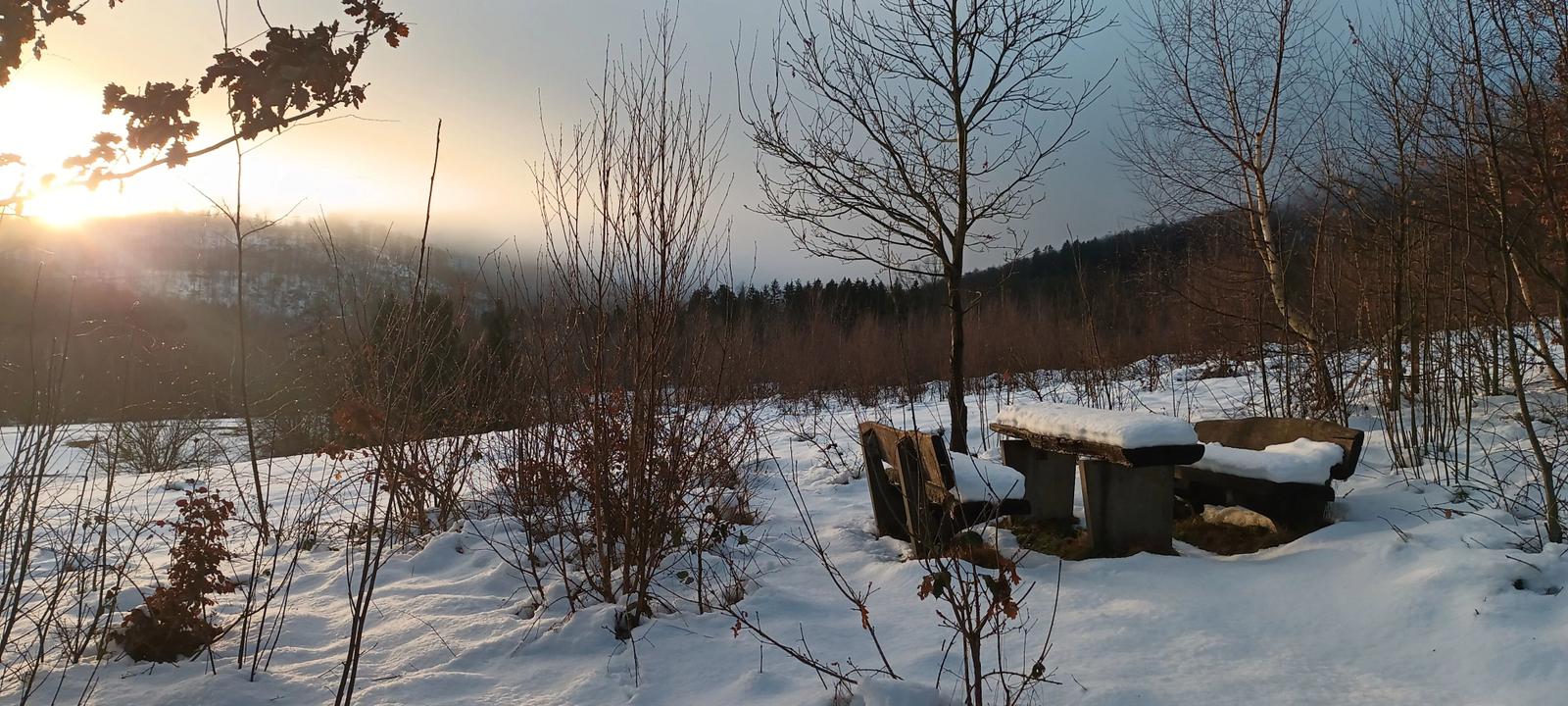 Schneebedeckter Holztisch und Bänke im Wald bei Sonnenaufgang.