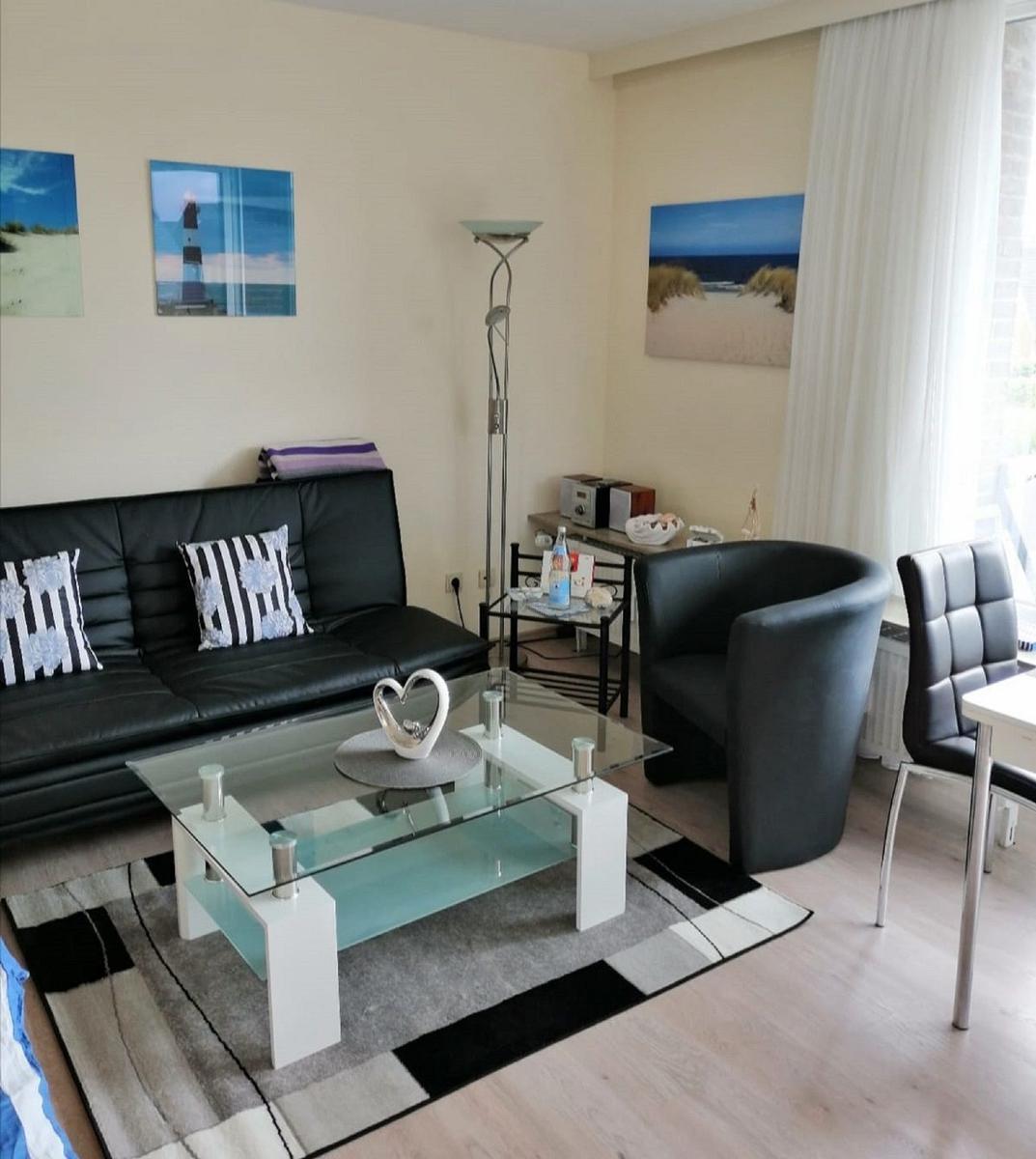 Wohnzimmer mit Sofa, Sessel und Glas-Tisch. Wandbilder zeigen Strand und Leuchtturm.