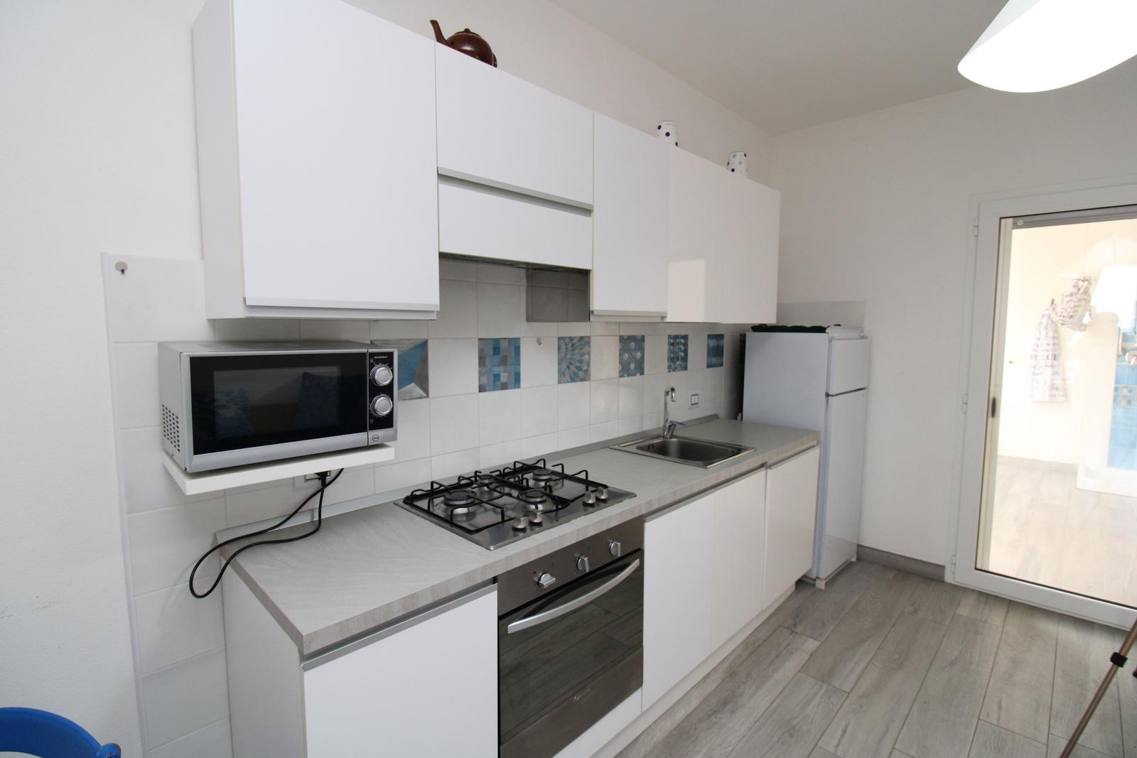 Küche mit weißen Schränken, Herd, Mikrowelle und Kühlschrank. Durchgang zur Terrasse.