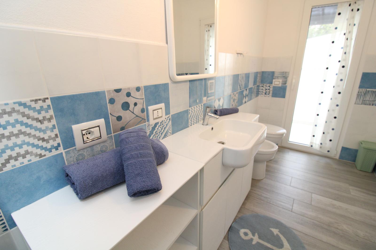 Badezimmer mit blau-weißen Fliesen, Waschbecken, Toilette und Badematte mit Ankern.