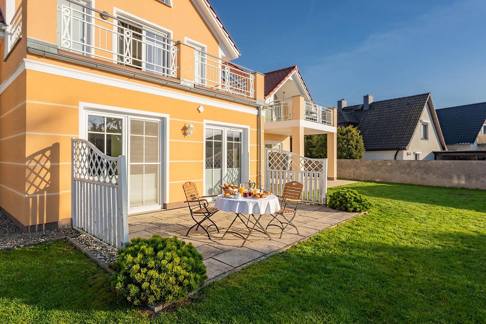 Haus mit Terrasse, Tisch und Stühlen im Garten. Weiße Gittertore und grünes Gras.