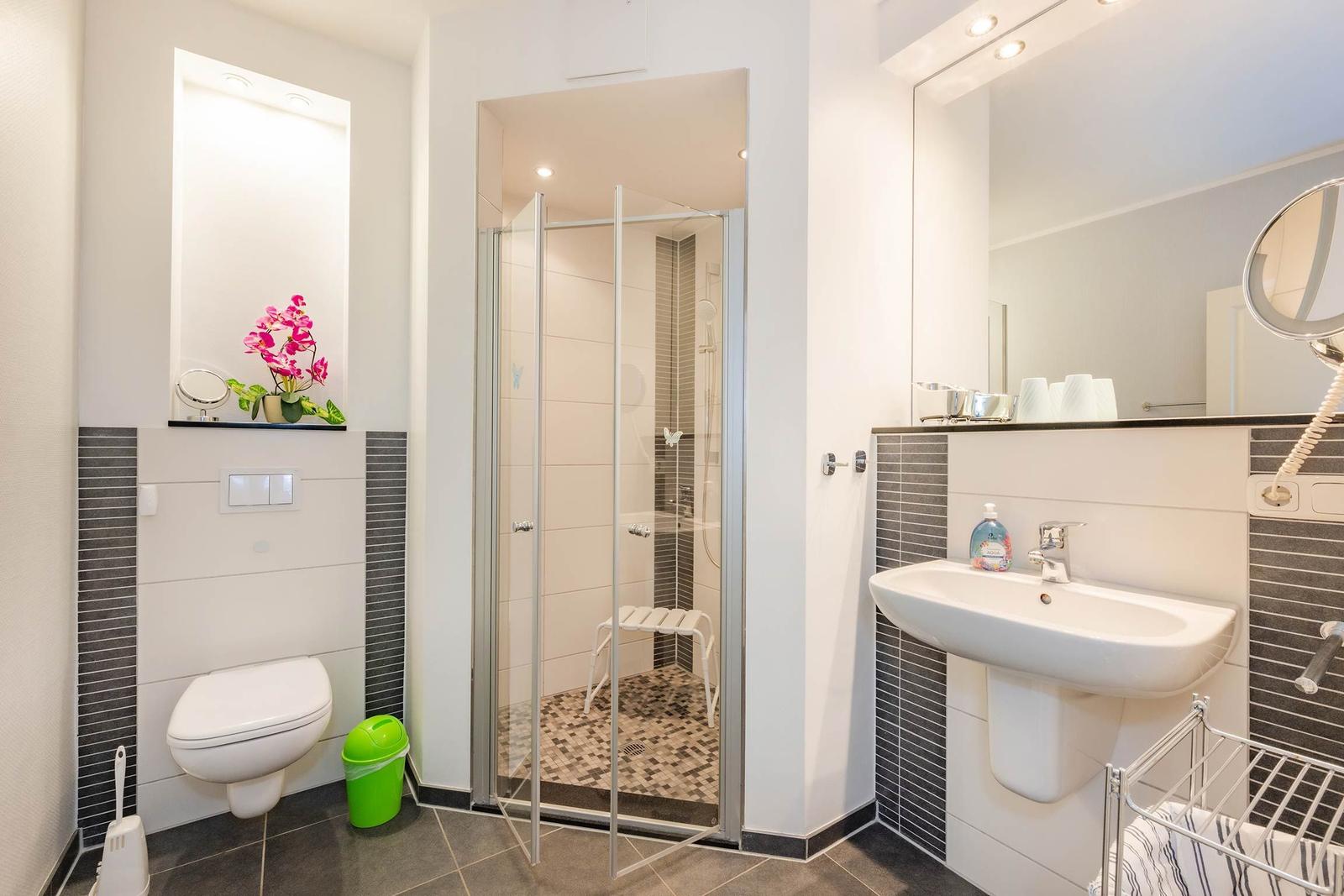 Badezimmer mit Dusche, Waschbecken und Toilette. Pinker Blumenstrauß auf Regal.