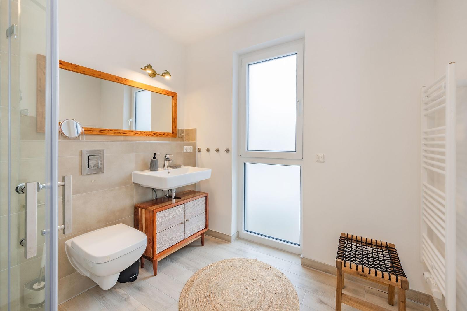 Badezimmer mit Dusche, Waschbecken und Toilette. Holzregal, Spiegel und Fenster sichtbar.