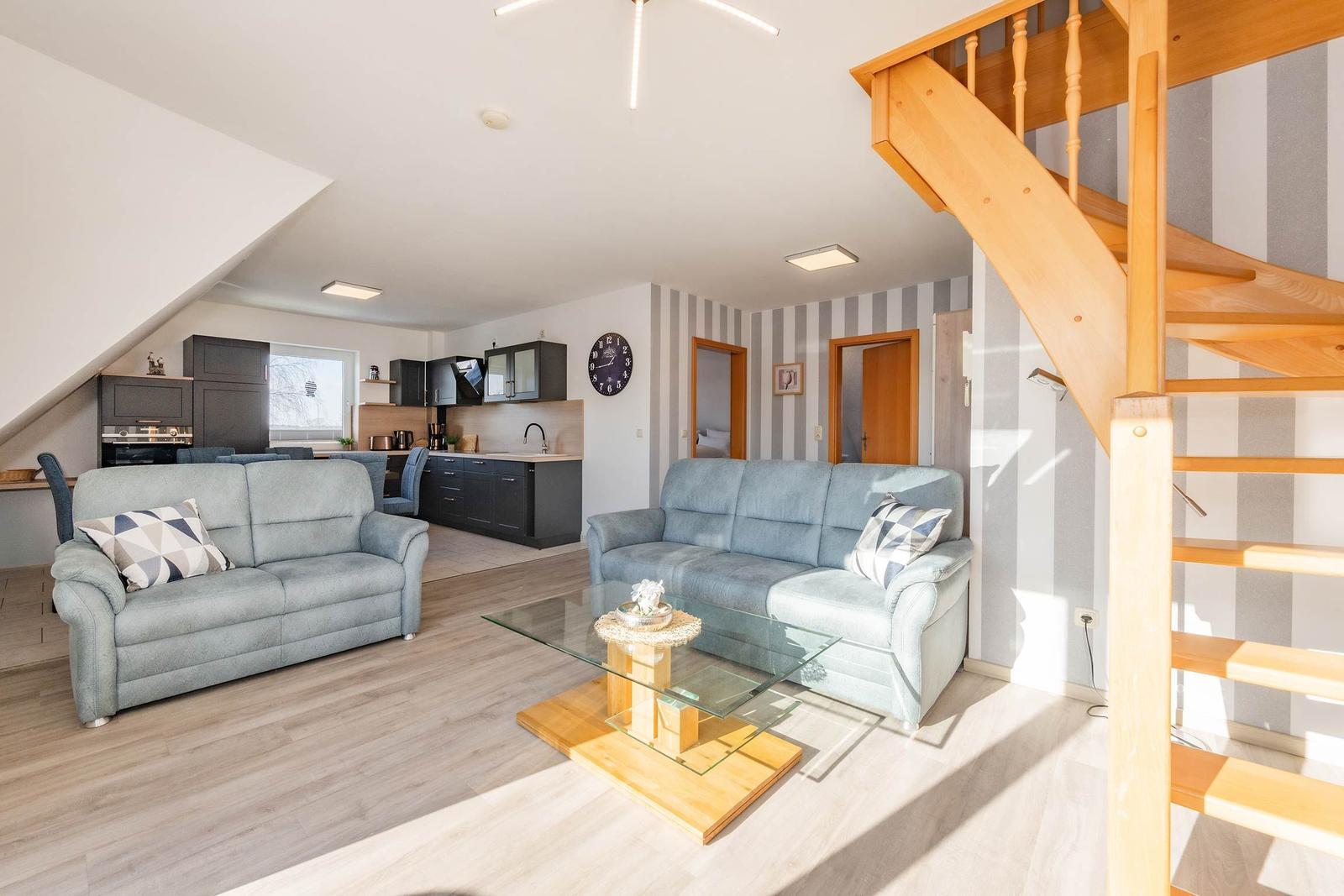 Wohn- und Küchenbereich mit zwei Sofas, Glas-Tisch und Holztreppe.
