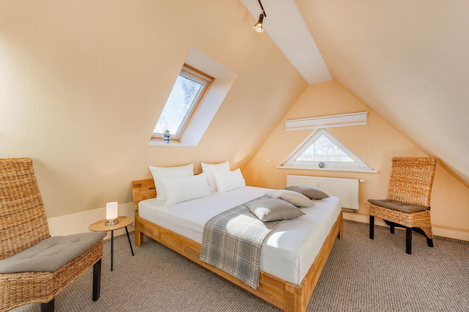 Schlafzimmer mit Dachschrägen, Bett, zwei Stühlen und zwei Fenstern.