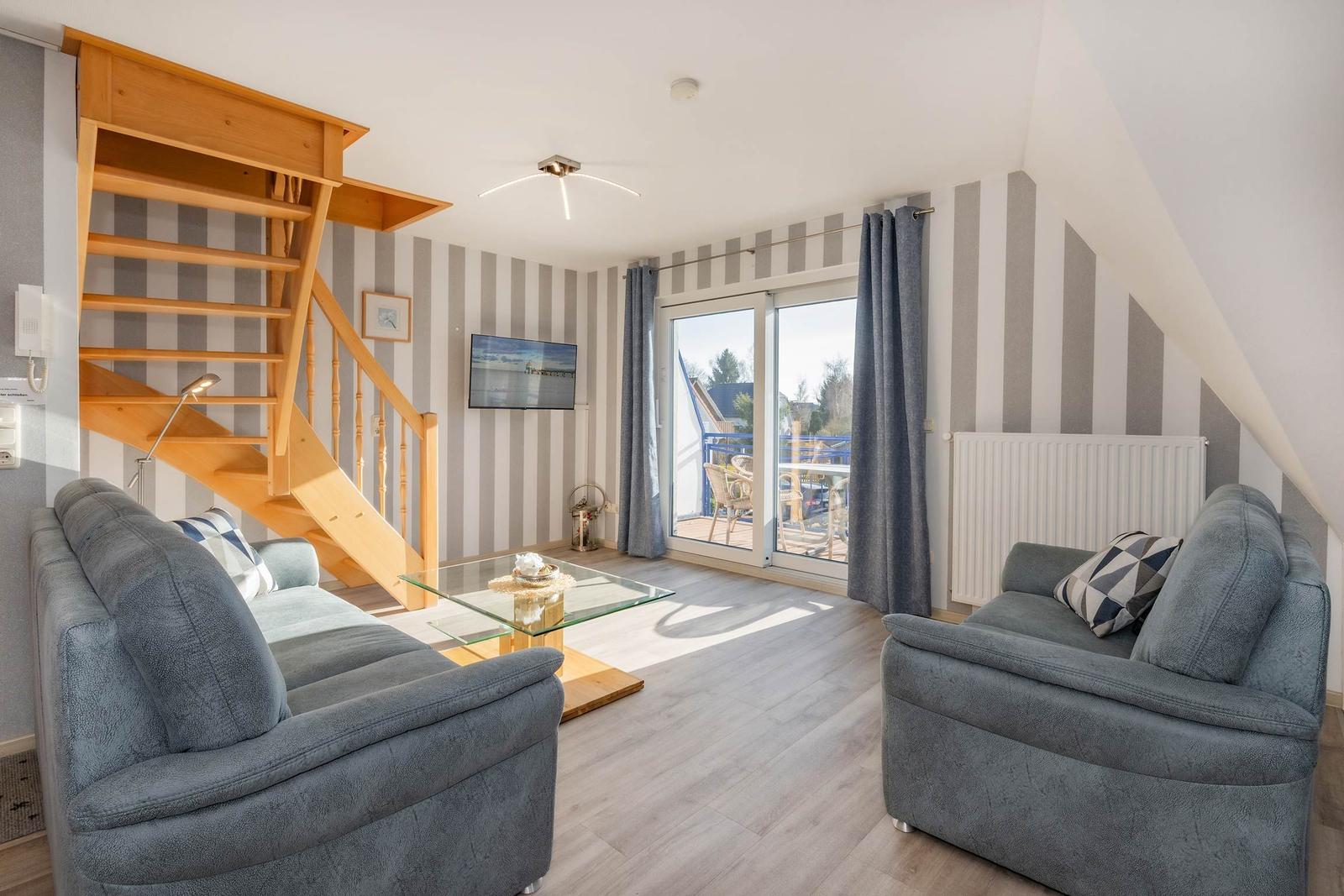 Wohnzimmer mit zwei Sofas, Glas-Tisch und Holztreppe. Balkon mit Blick nach draußen.