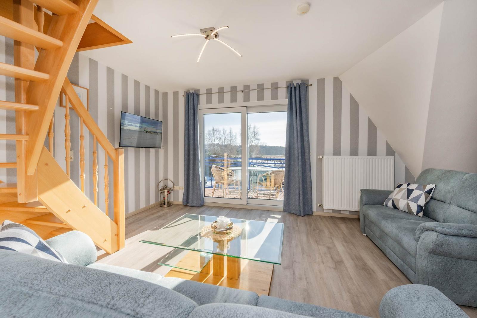 Wohnzimmer mit Sofa, Glas-Tisch und Balkonansicht. Holztreppe und gestreifte Wände.