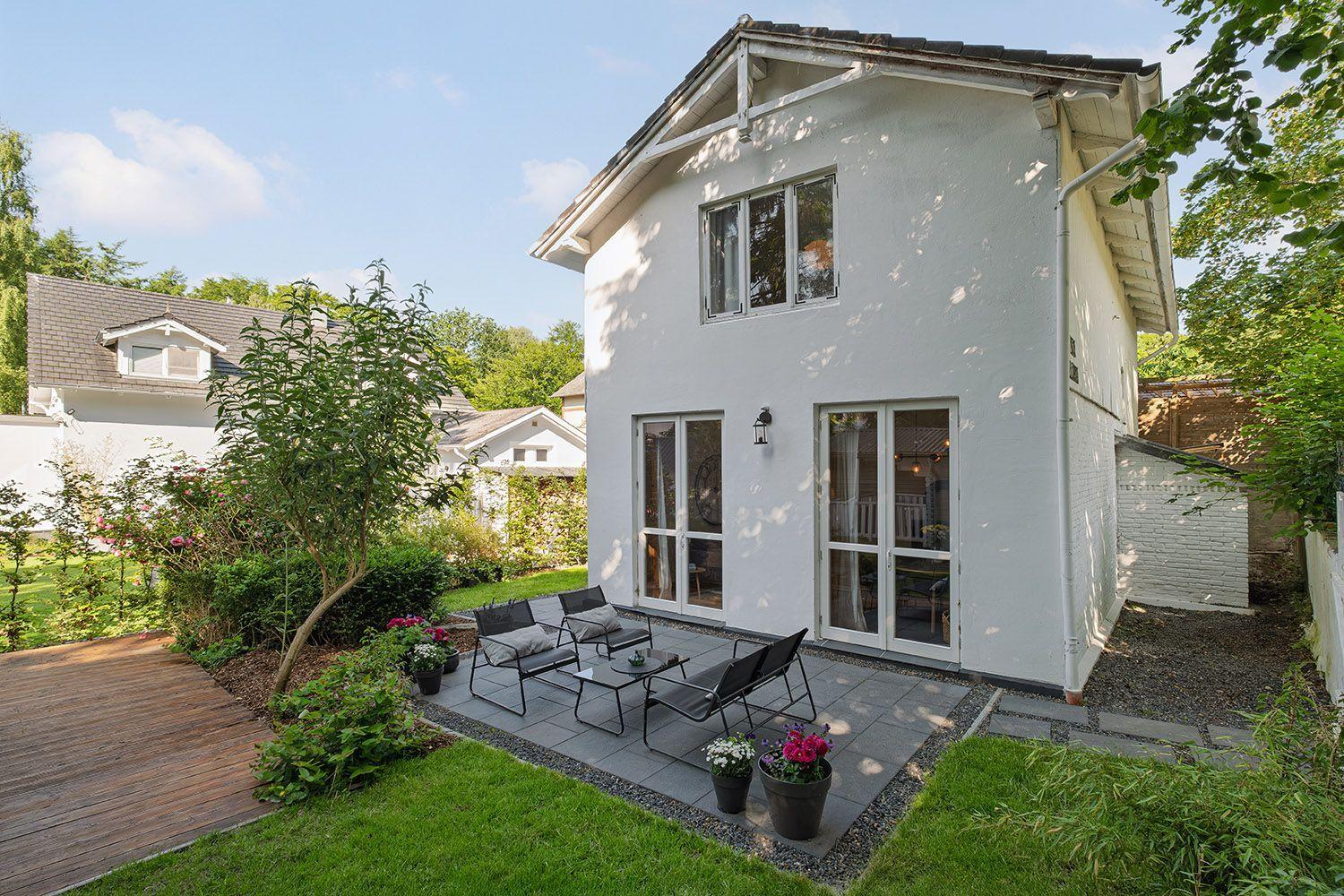 Weiße Villa mit Terrasse und Garten. Glastüren führen ins Haus. Möbel und Pflanzen sind auf der Terrasse.