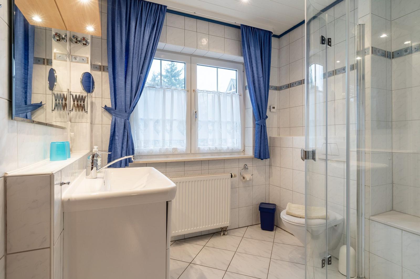 Badezimmer mit Dusche, Waschbecken und Fenster mit Vorhängen.