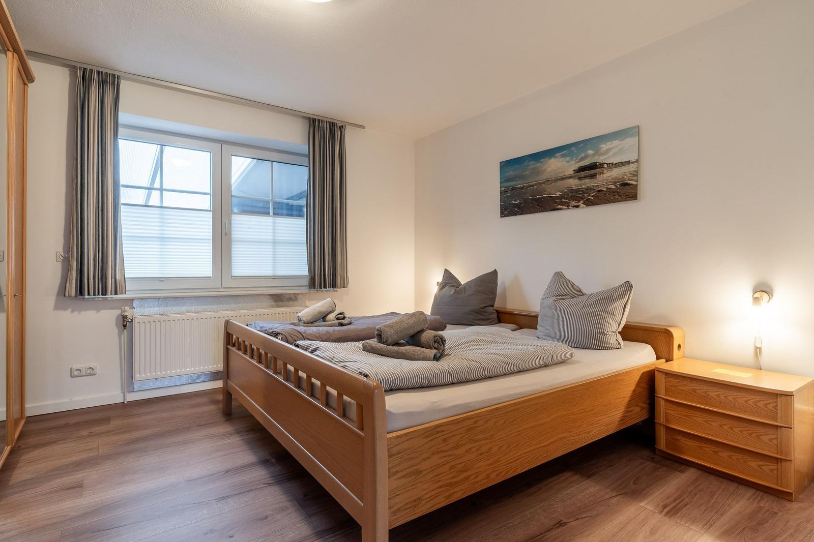 Schlafzimmer mit Holzbett, Nachttisch und Fenster mit Vorhängen.