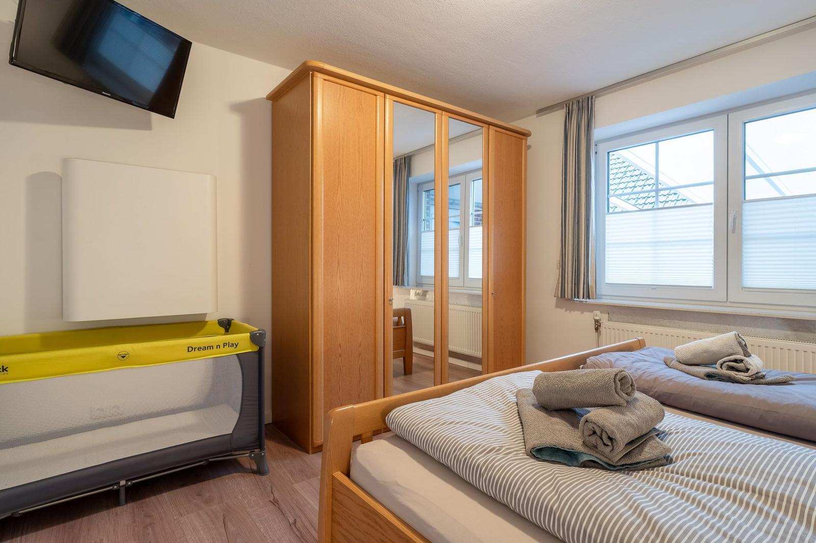 Zimmer mit Bett, Kleiderschrank und Babybett. Fenster mit Jalousien und Wand-TV.