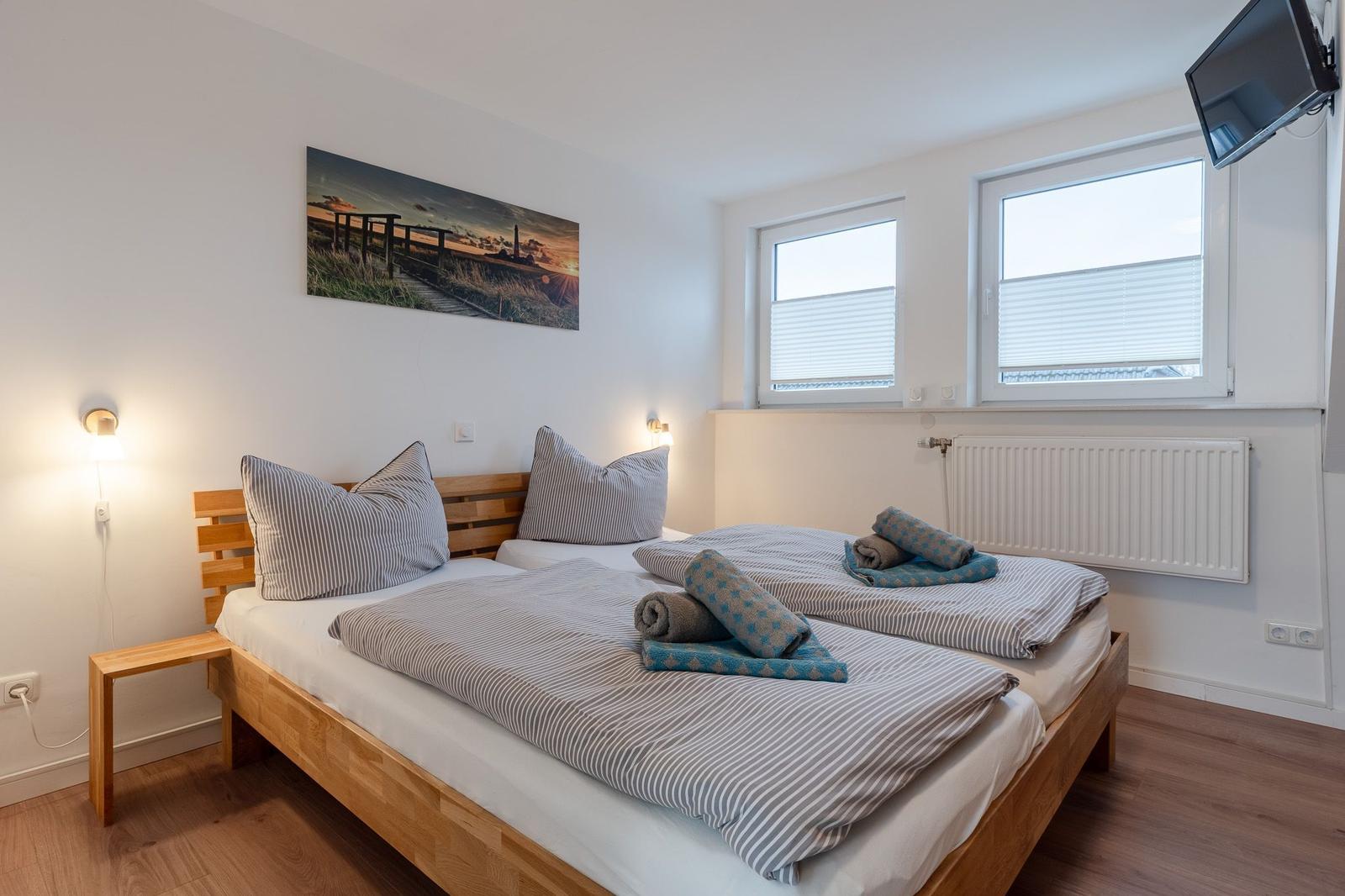 Schlafzimmer mit Doppelbett, Wandbild, zwei Fenstern und TV.
