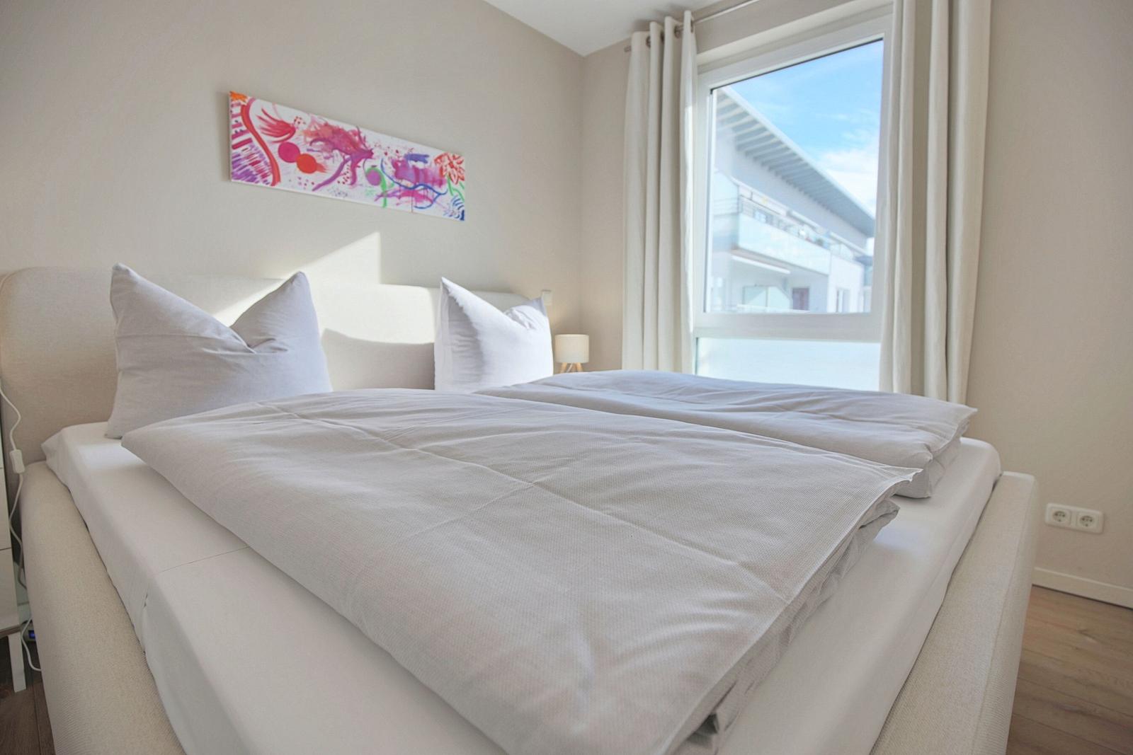 Ein Bett mit weißen Kissen und Bettwäsche. An der Wand hängt ein buntes Bild. Ein Fenster mit weißen Vorhängen.