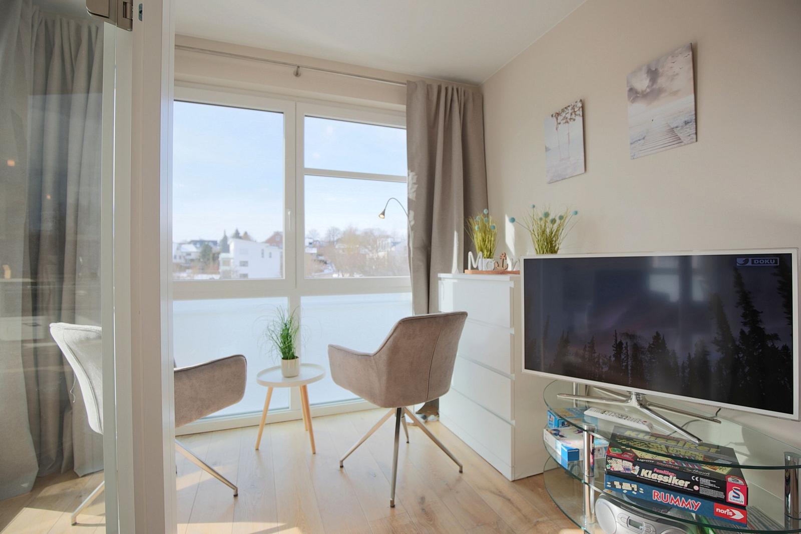 Wohnraum mit TV, Schrank und Sitzgelegenheit vor großem Fenster.