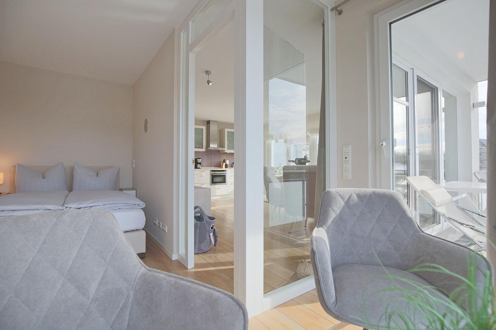 Schlafzimmer mit Blick auf Küche und Balkon. Zwei Sessel und Bett sind sichtbar.
