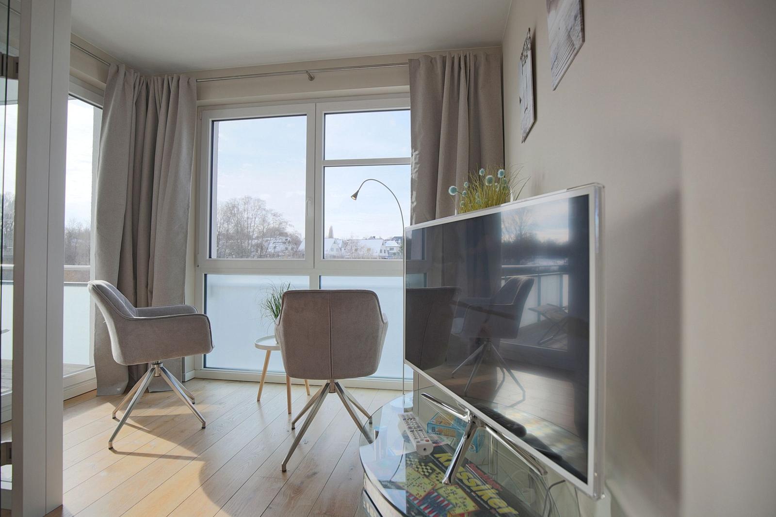 Wohnzimmer mit TV, zwei Stühlen und großem Fenster mit Blick auf Außenbereich.