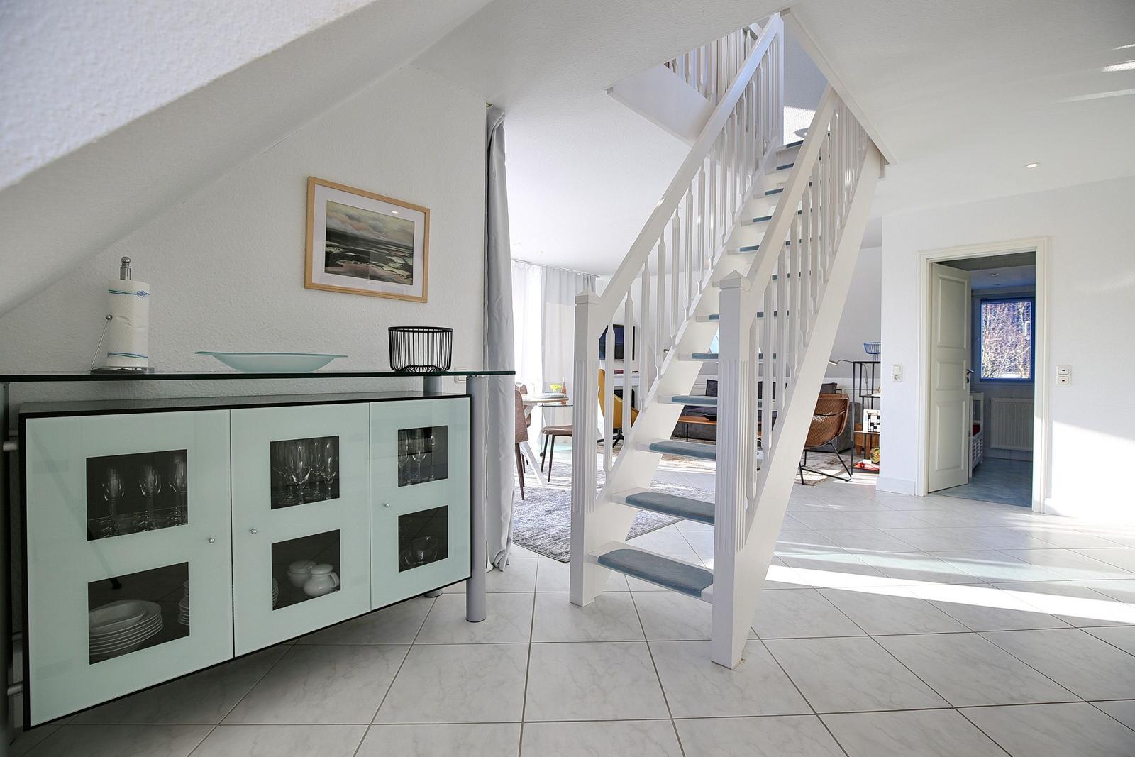 Eingangsbereich mit Treppenhaus und Glasvitrine. Weiße Wände und Fliesenboden.