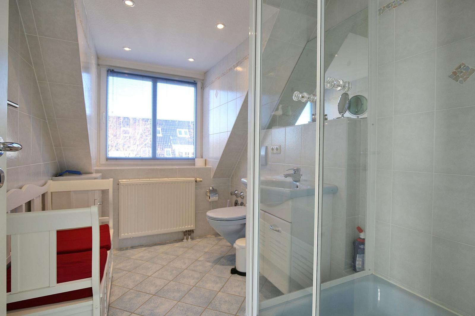 Badezimmer mit Dusche, Toilette und Fenster. Ein weißer Kinderstuhl mit rotem Kissen steht daneben.