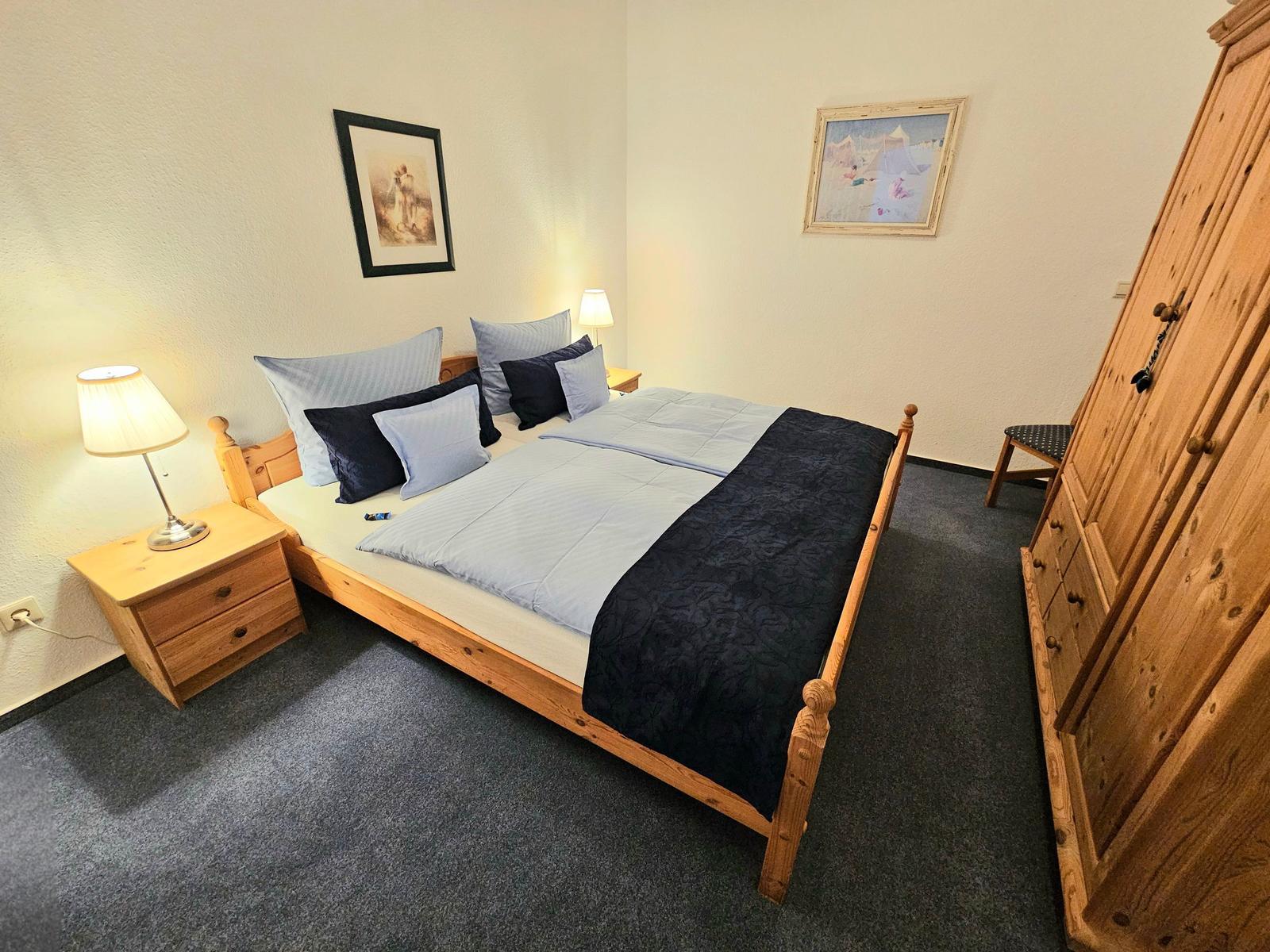 Schlafzimmer mit Holzbett, Nachttisch, Kleiderschrank und Wandbildern.
