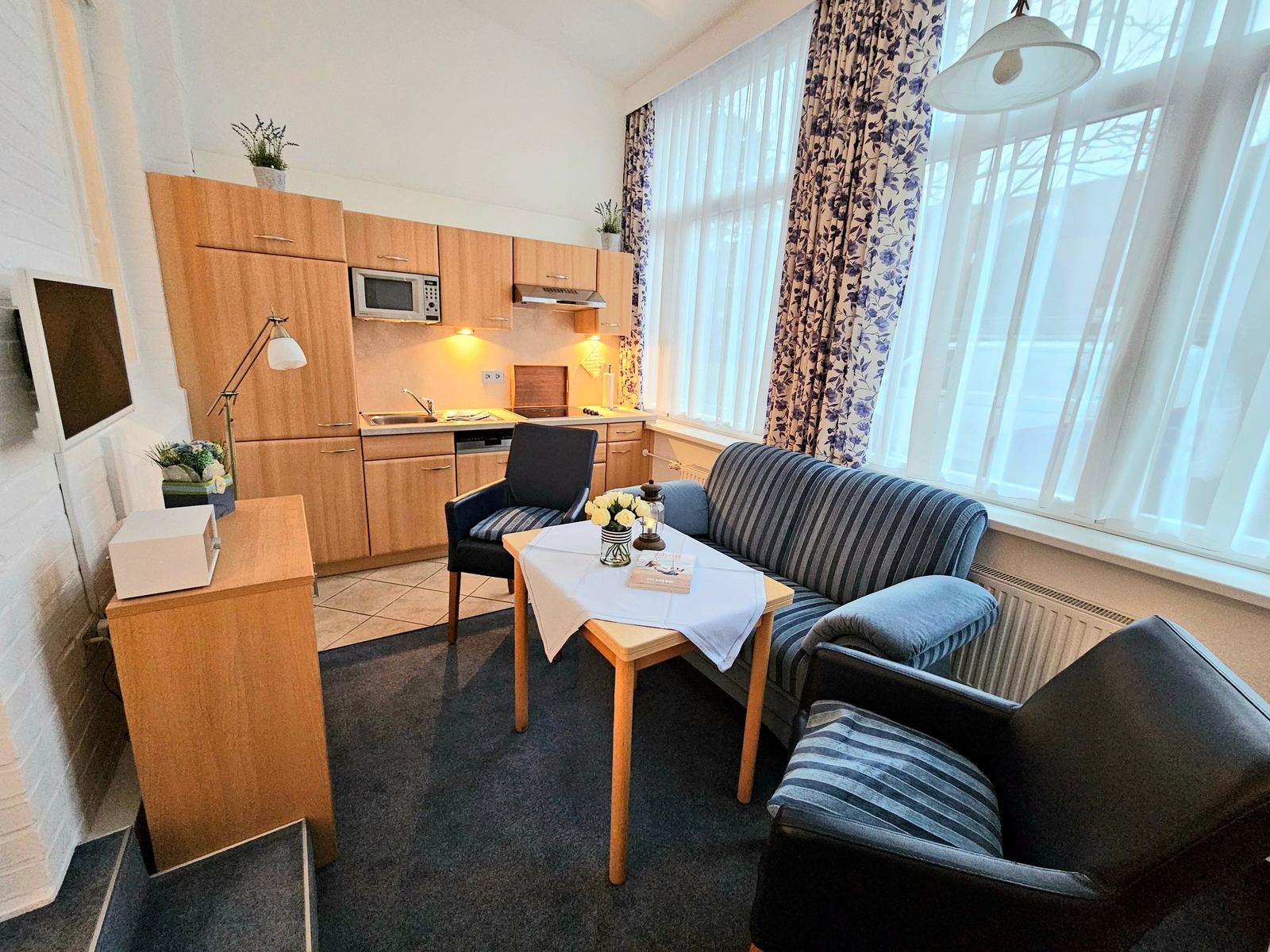 Wohnbereich mit Sofa, Tisch, Küchenzeile und Fenster mit Vorhängen.