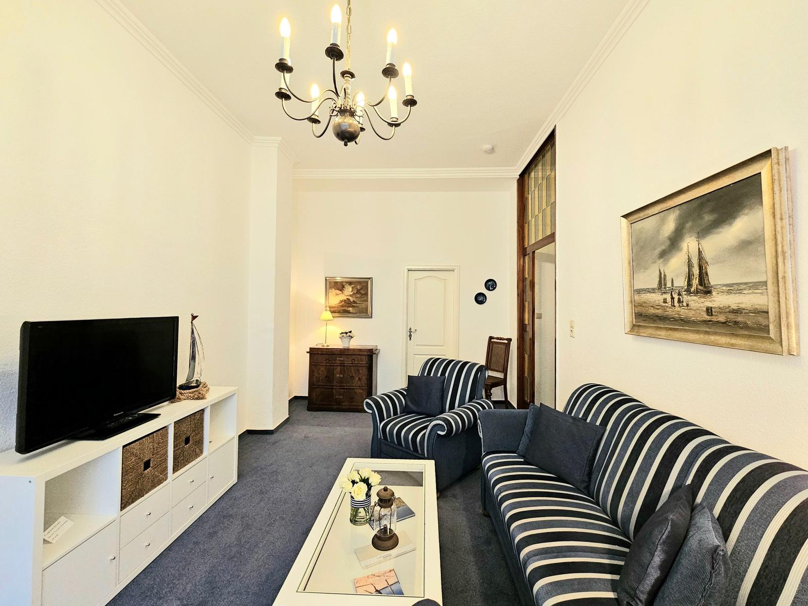 Wohnzimmer mit Sofa, Sessel und Fernseher. Deckenleuchte und Wandbild sichtbar.