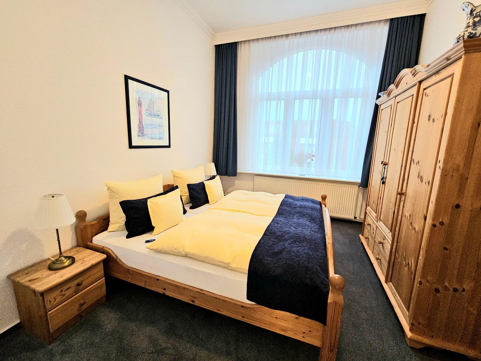 Schlafzimmer mit Holzbett, Kleiderschrank und Fenster mit Vorhängen.