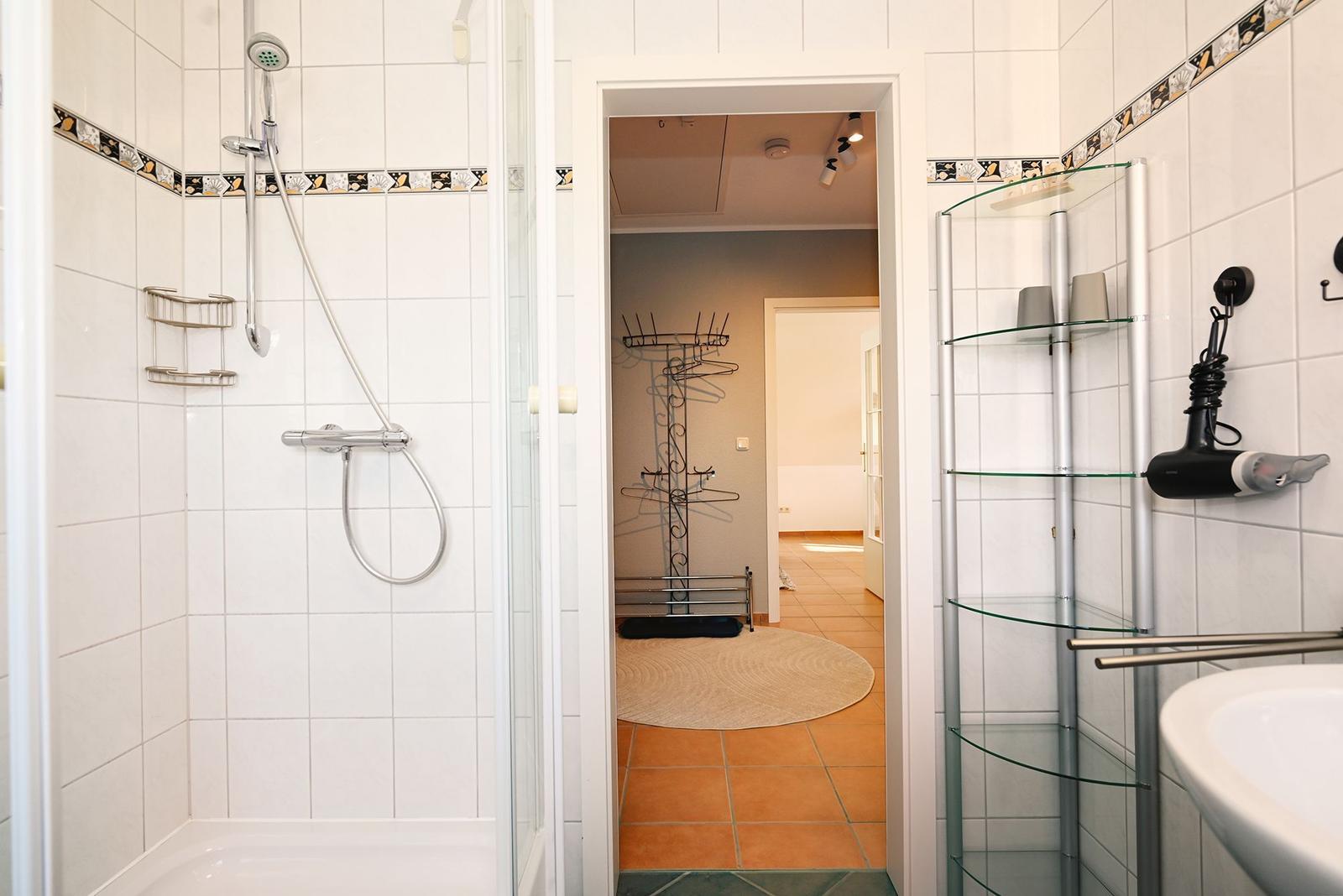 Badezimmer mit Dusche, Waschbecken und Glasregal. Durchgang zu einem Raum mit Kleiderständer.