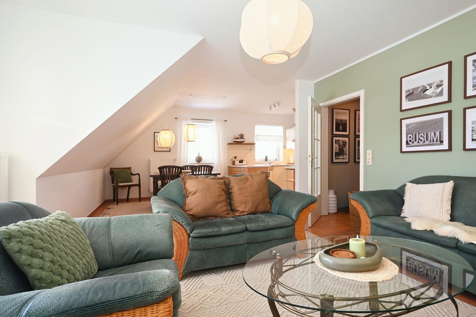 Wohnzimmer mit grünen Sofas, Glas-Tisch und Wandbildern. Küchenbereich im Hintergrund.