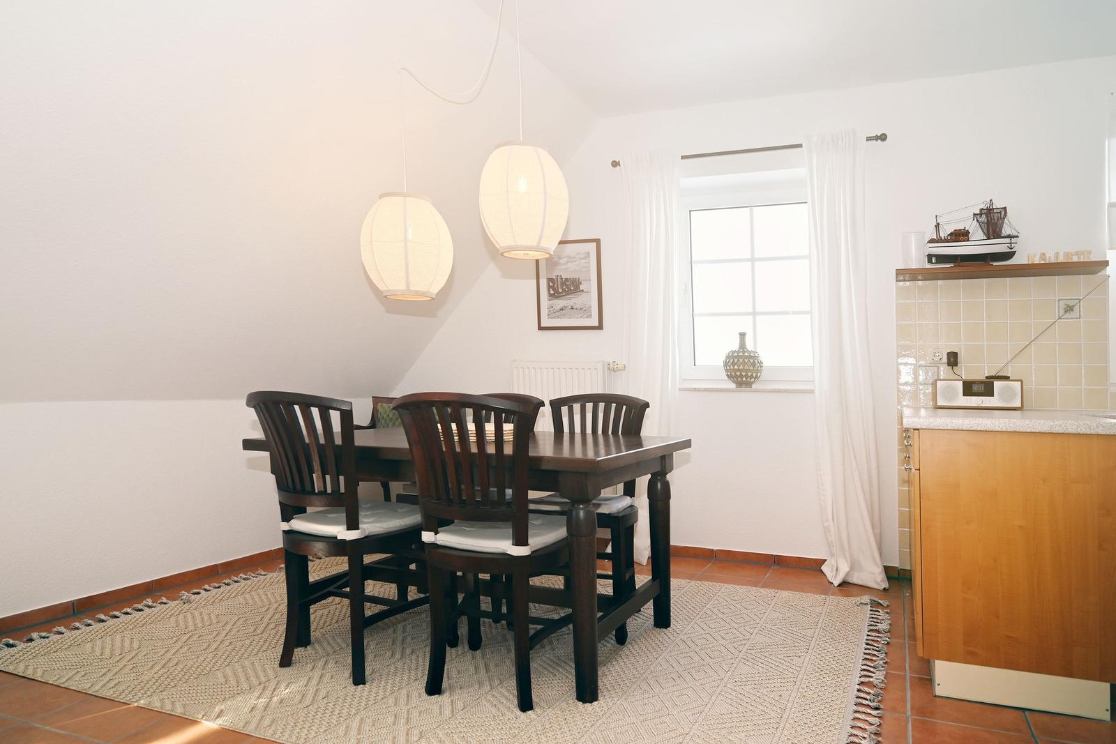Esszimmer mit Holztisch, Stühlen und zwei Hängelampen. Fenster mit Vorhängen und Küchenzeile.