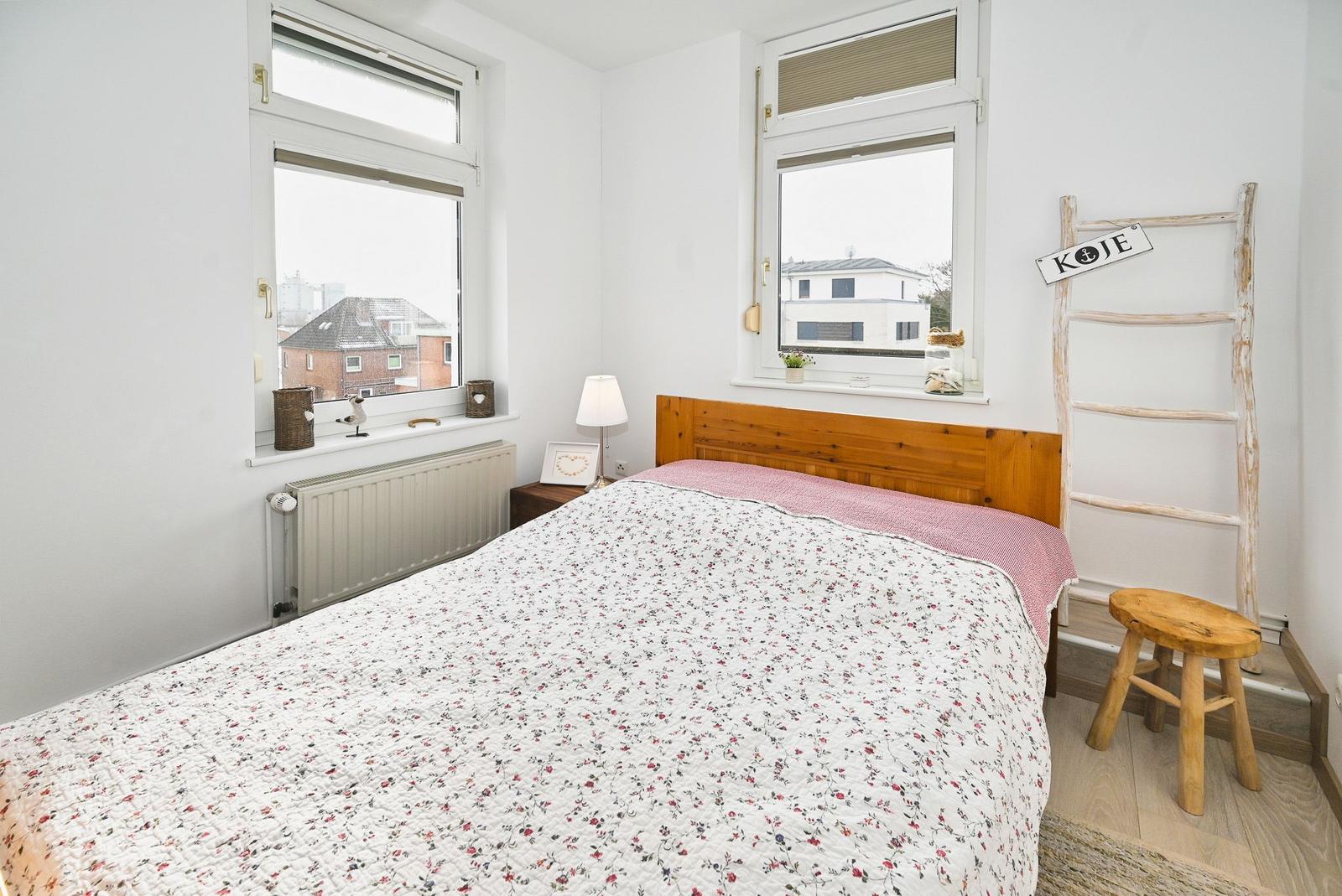 Schlafzimmer mit Bett, zwei Fenstern und Holzregal.