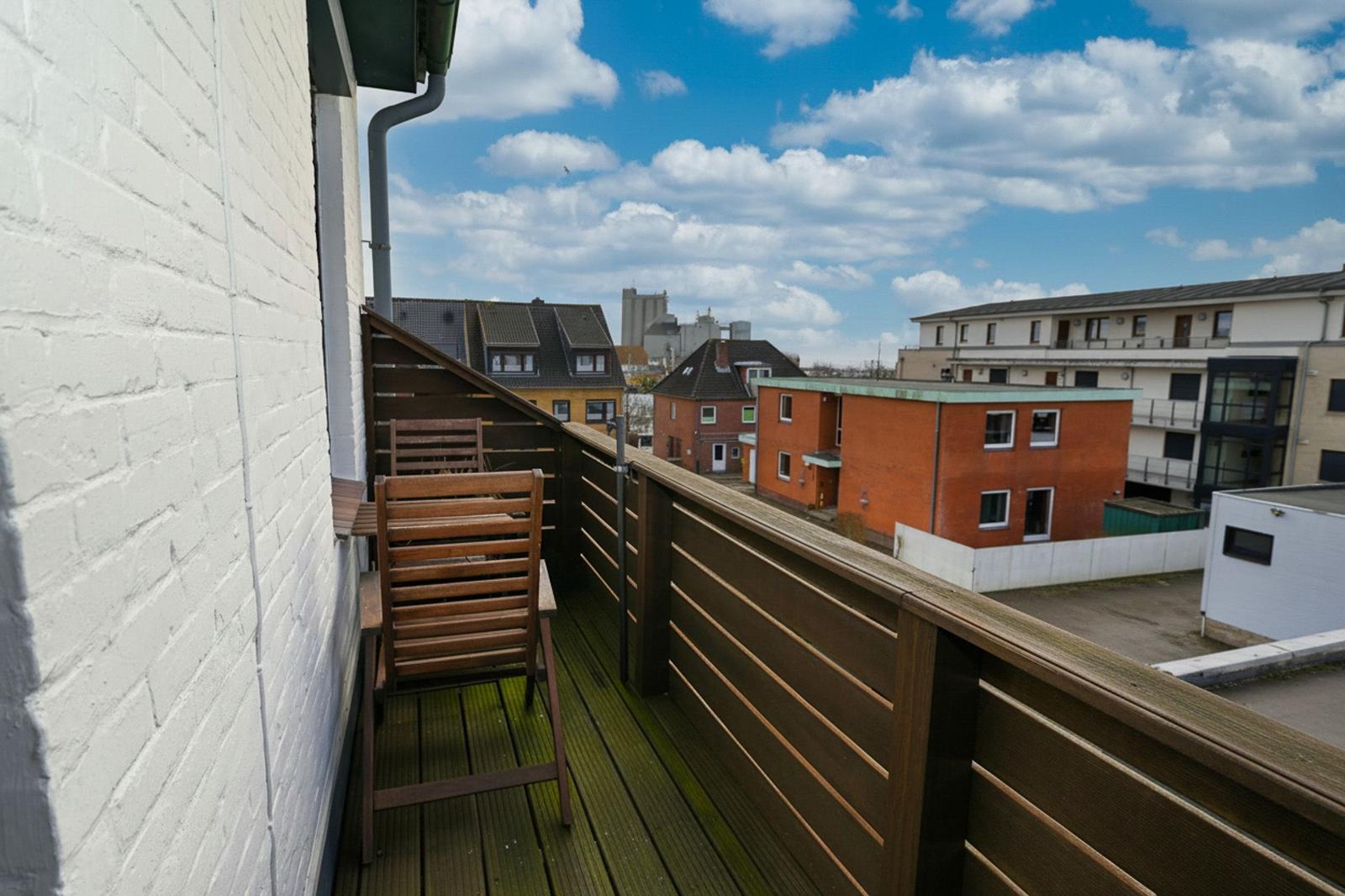 Terrasse mit Holzgeländer und Stuhl. Blick auf Wohnhäuser und Himmel.