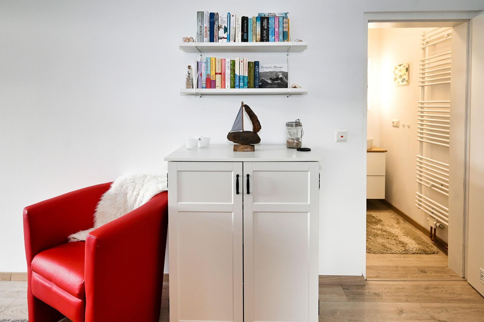 Roter Sessel neben weißem Schrank mit Bücherregal darüber. Blick in angrenzenden Raum.