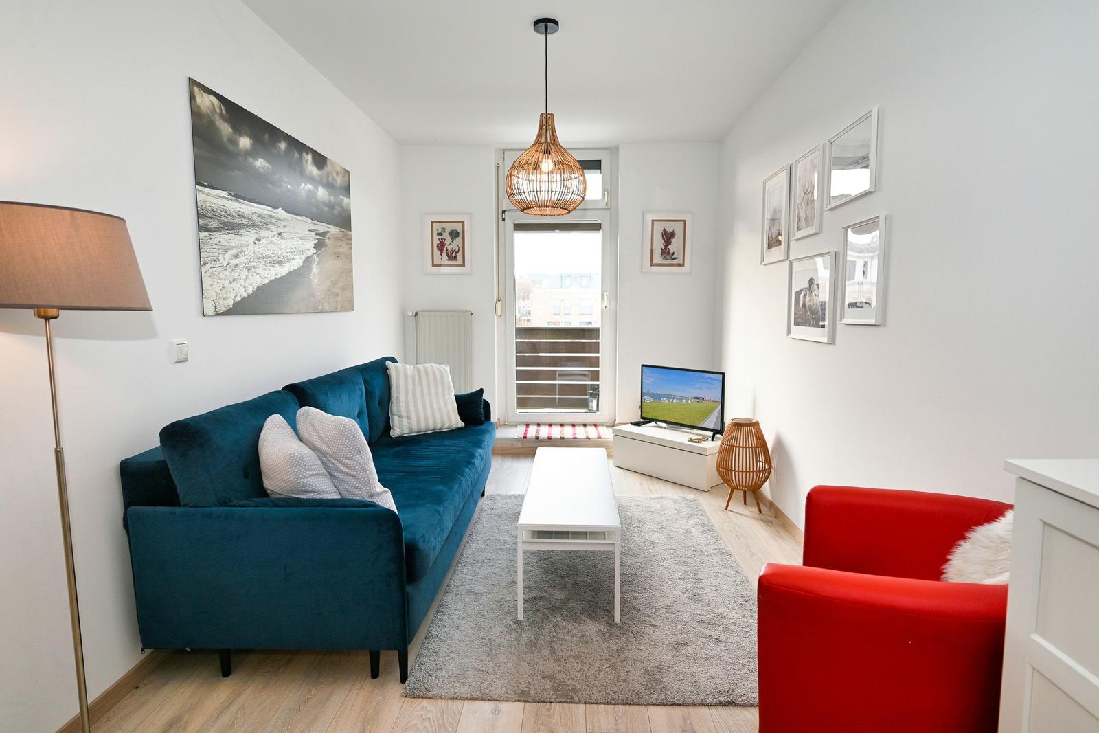 Wohnzimmer mit blauem Sofa, rotem Sessel und Fernseher. Helle Wände und Holzboden.