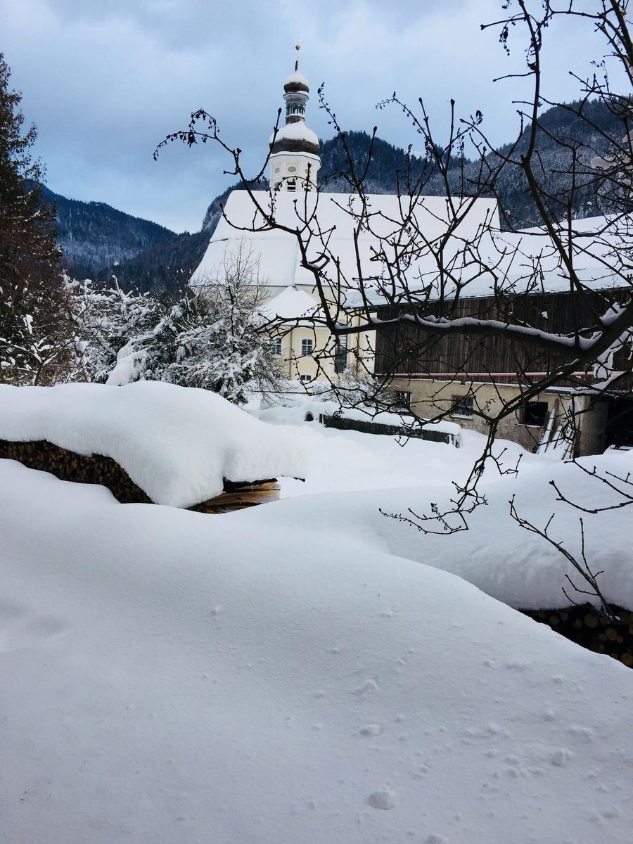 Snow-covered church with tower in winter, surrounded by mountains and bare branches.