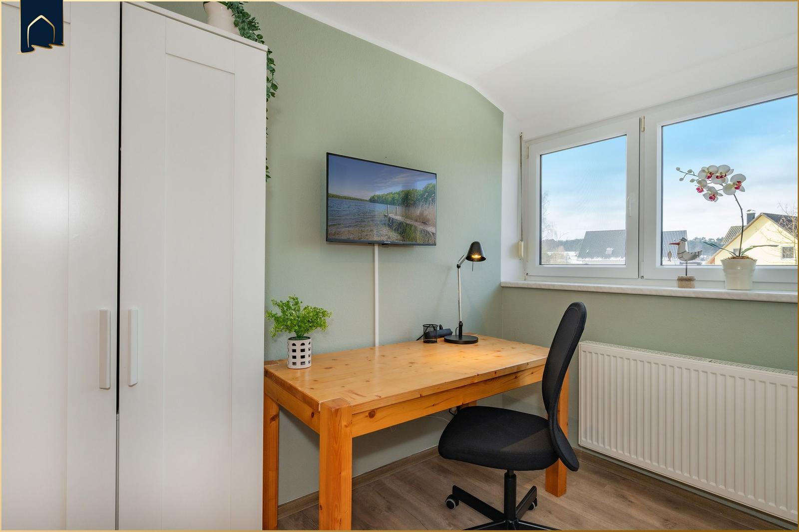 Ein Arbeitszimmer mit Schreibtisch, Stuhl und Fernseher an der Wand. Fenster mit Blick nach draußen.