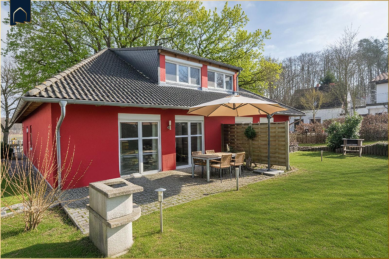 Rotes Haus mit Terrasse, Garten und Sitzgruppe unter Sonnenschirm.