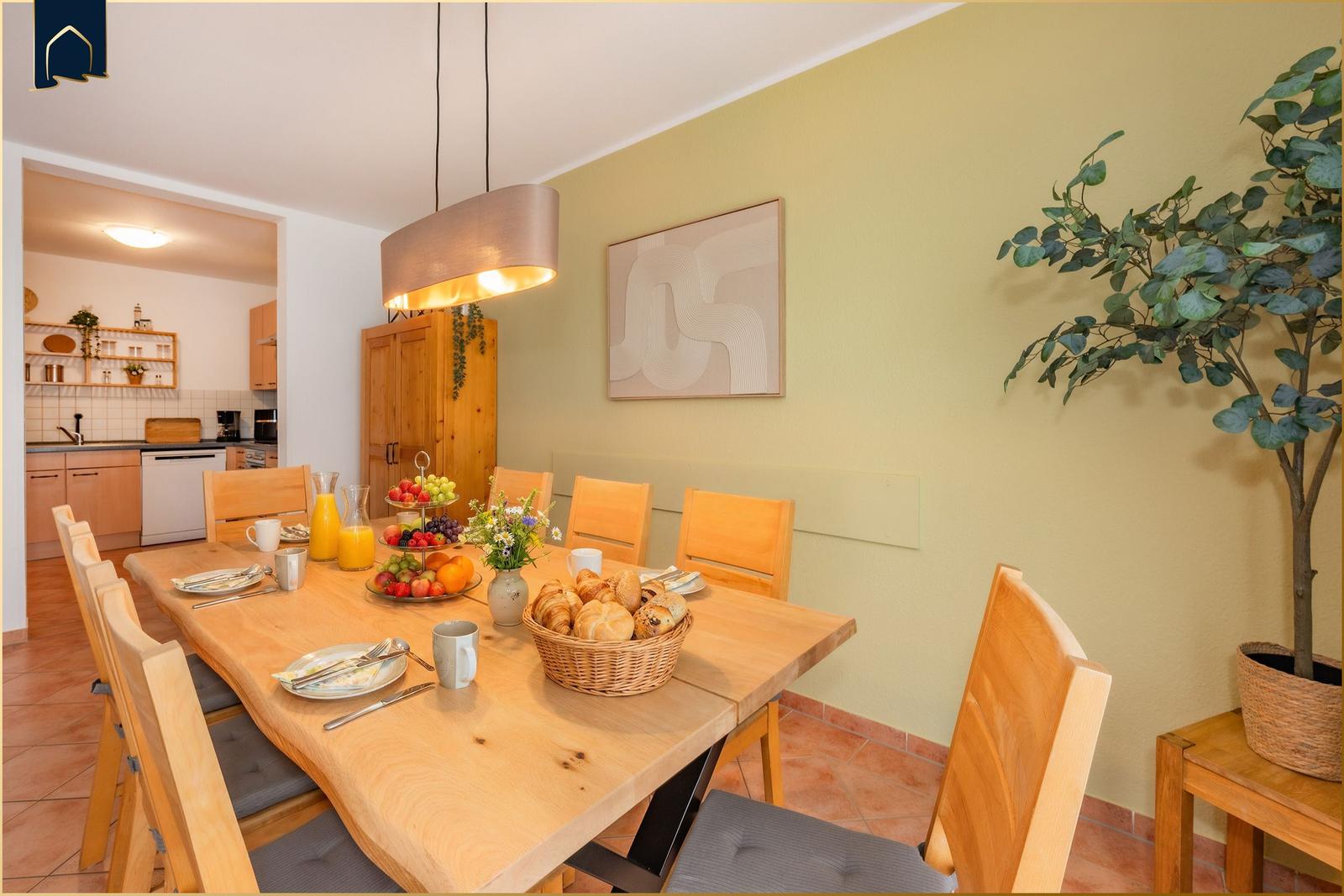 Essbereich mit Holztisch, Stühlen und Obst. Küche im Hintergrund. Wandbild und Zimmerpflanze.