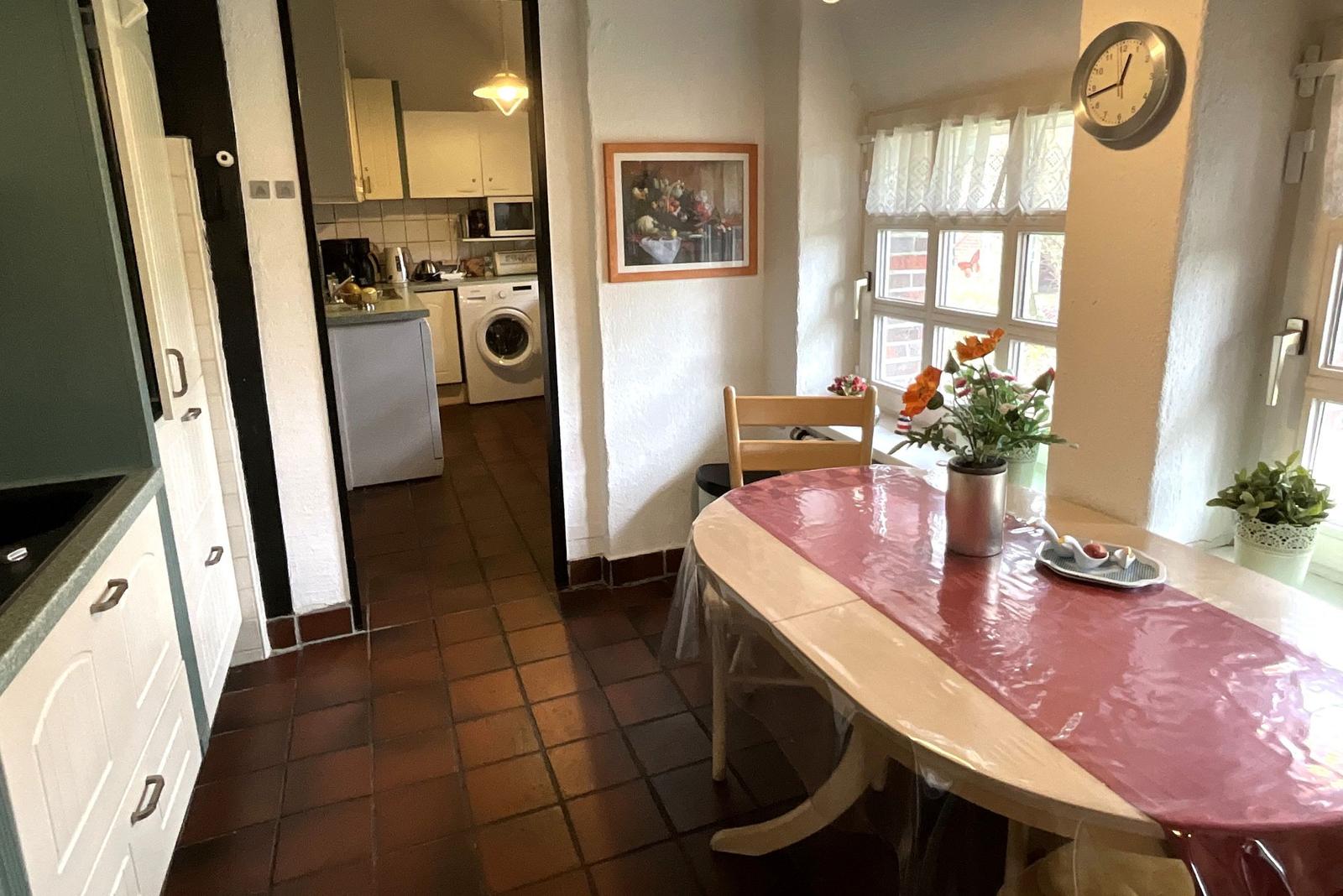 Essbereich mit Tisch, Stühlen und Fenster. Küche mit Waschmaschine sichtbar.