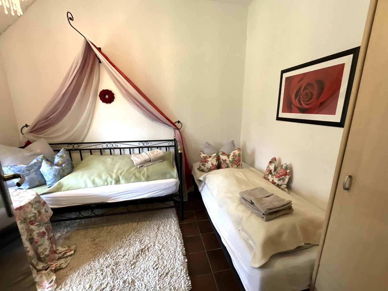 Zimmer mit Doppelbett und Einzelbett, Wandbild mit Rose, Teppich und Kleiderstange.