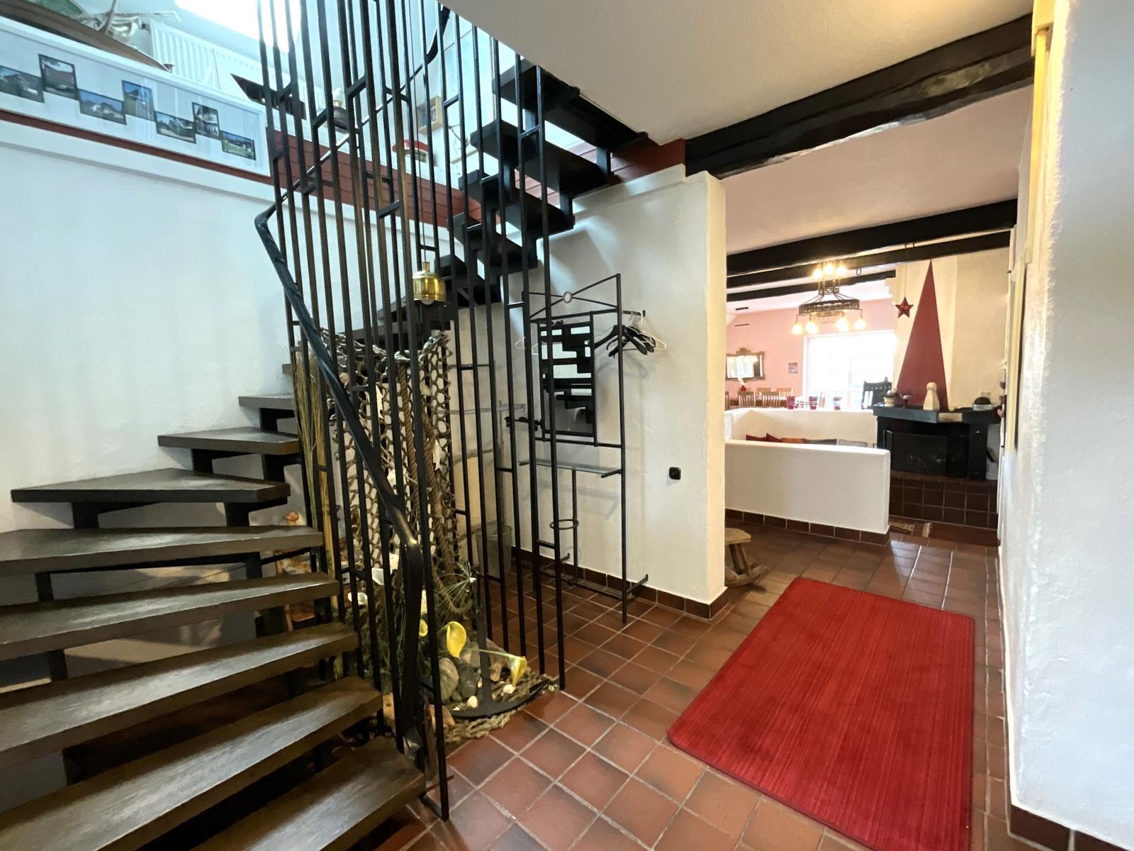 Eingangsbereich mit Treppe und rotem Teppich. Wohnbereich mit Kamin und Holzbalken sichtbar.