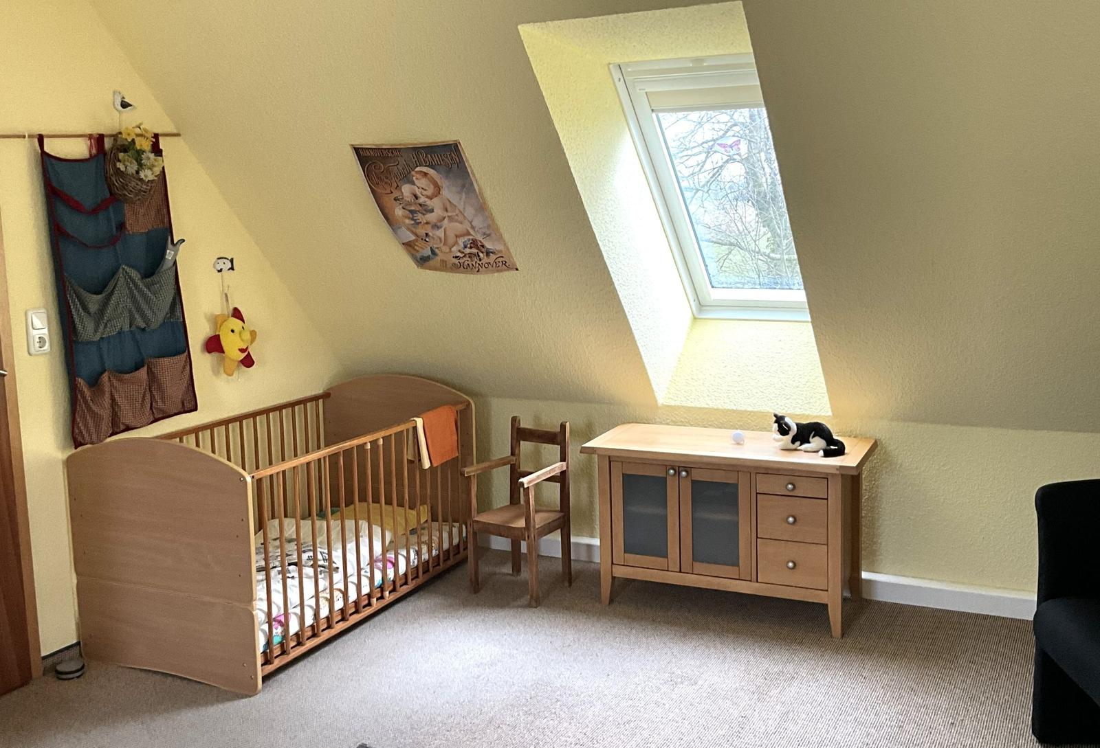 Kinderzimmer mit Babybett, Stuhl und Schrank unter Dachschräge mit Fenster.