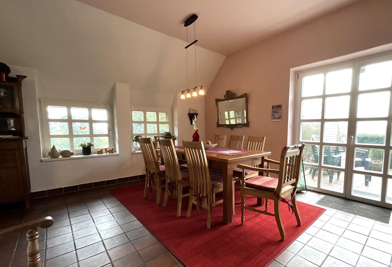 Esszimmer mit Holztisch, Stühlen und rotem Teppich. Fenster und Glastür führen nach draußen.