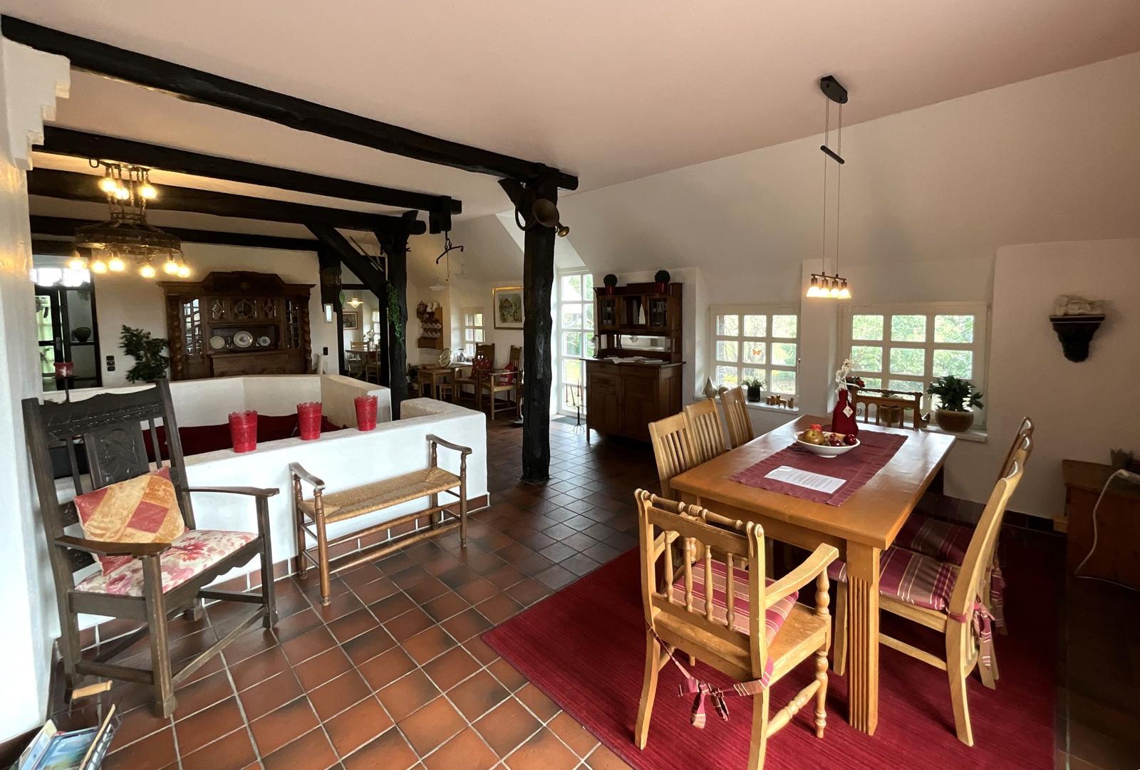 Großes Esszimmer mit Holztisch, Stühlen und Fenstern. Im Hintergrund Wohnbereich mit Holzbalken.