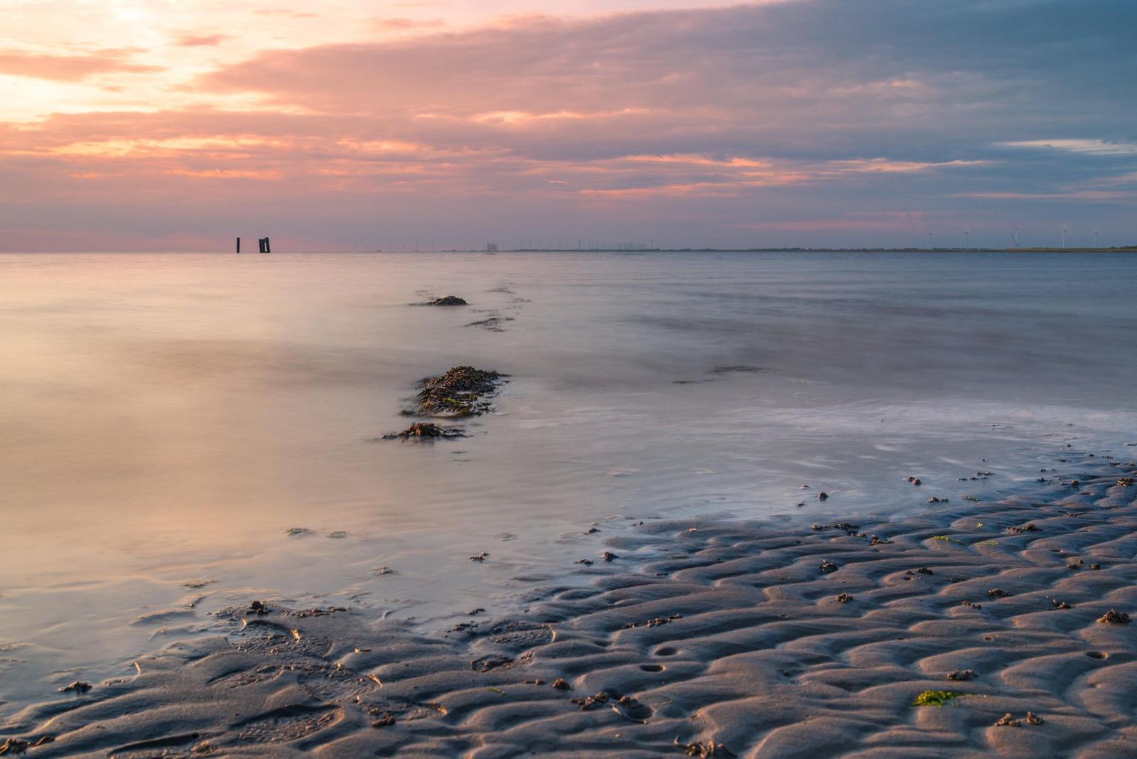 Sonnenuntergang über ruhigem Wasser mit Sandstrand und entfernten Windrädern.