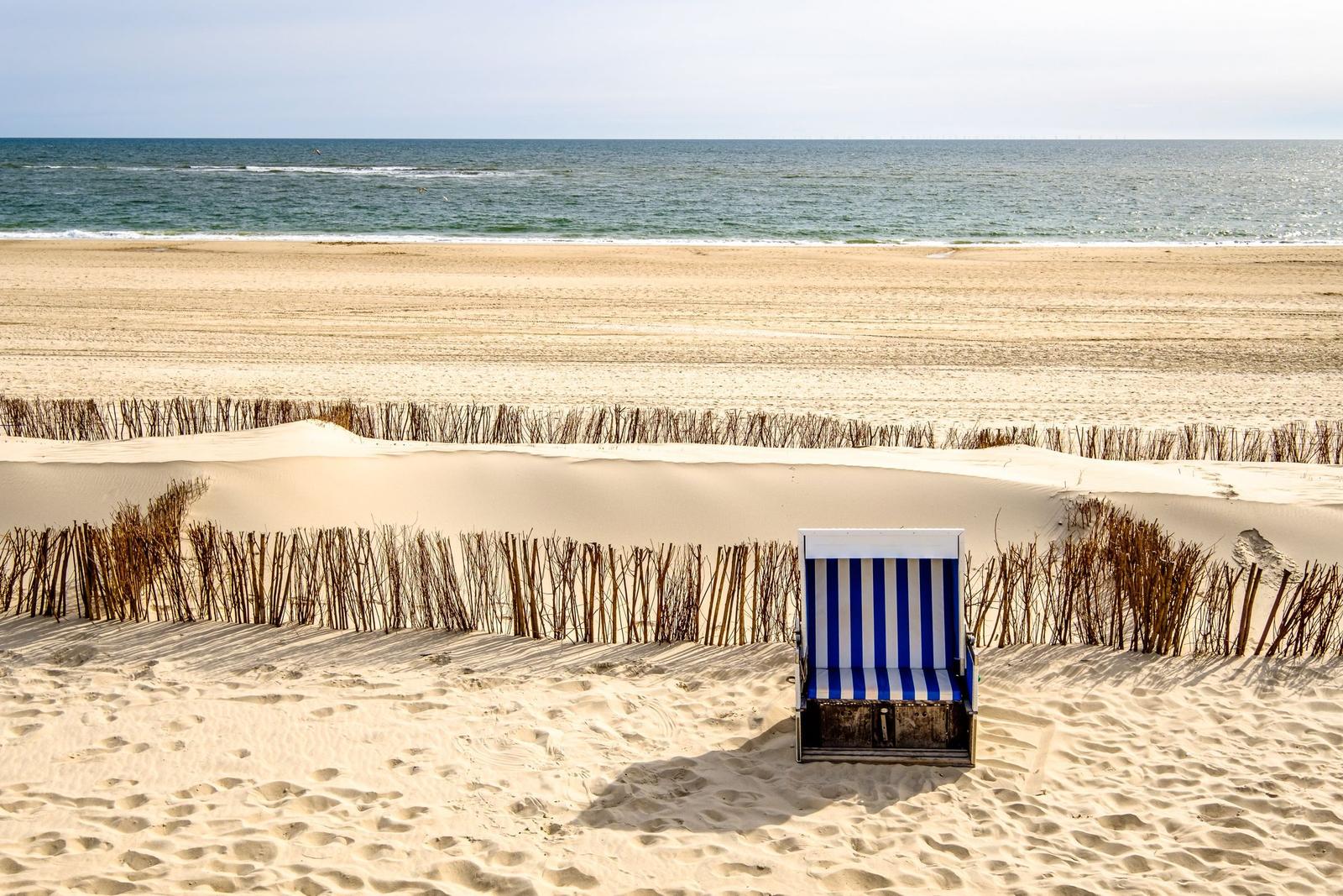Ein blau-weiß gestreifter Strandkorb auf Sand, hinter ihm Dünen und das Meer.